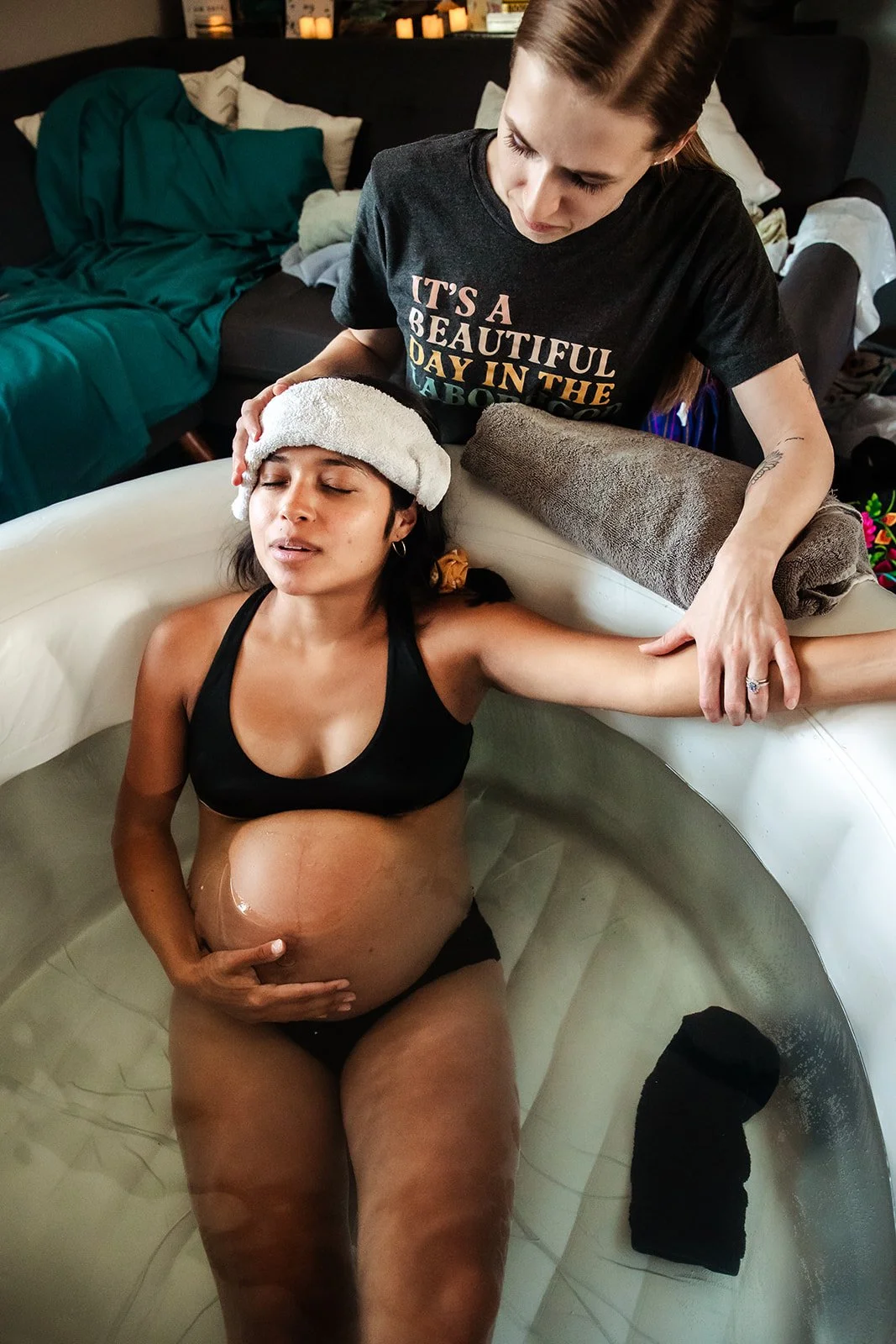 A woman with a towel wrapped on her head soaking in a bathtub, with a pregnant belly and a black sports bra, while another woman standing beside the tub holds her arm, wearing a black T-shirt with colorful text, and resting a towel on the edge of the tub.