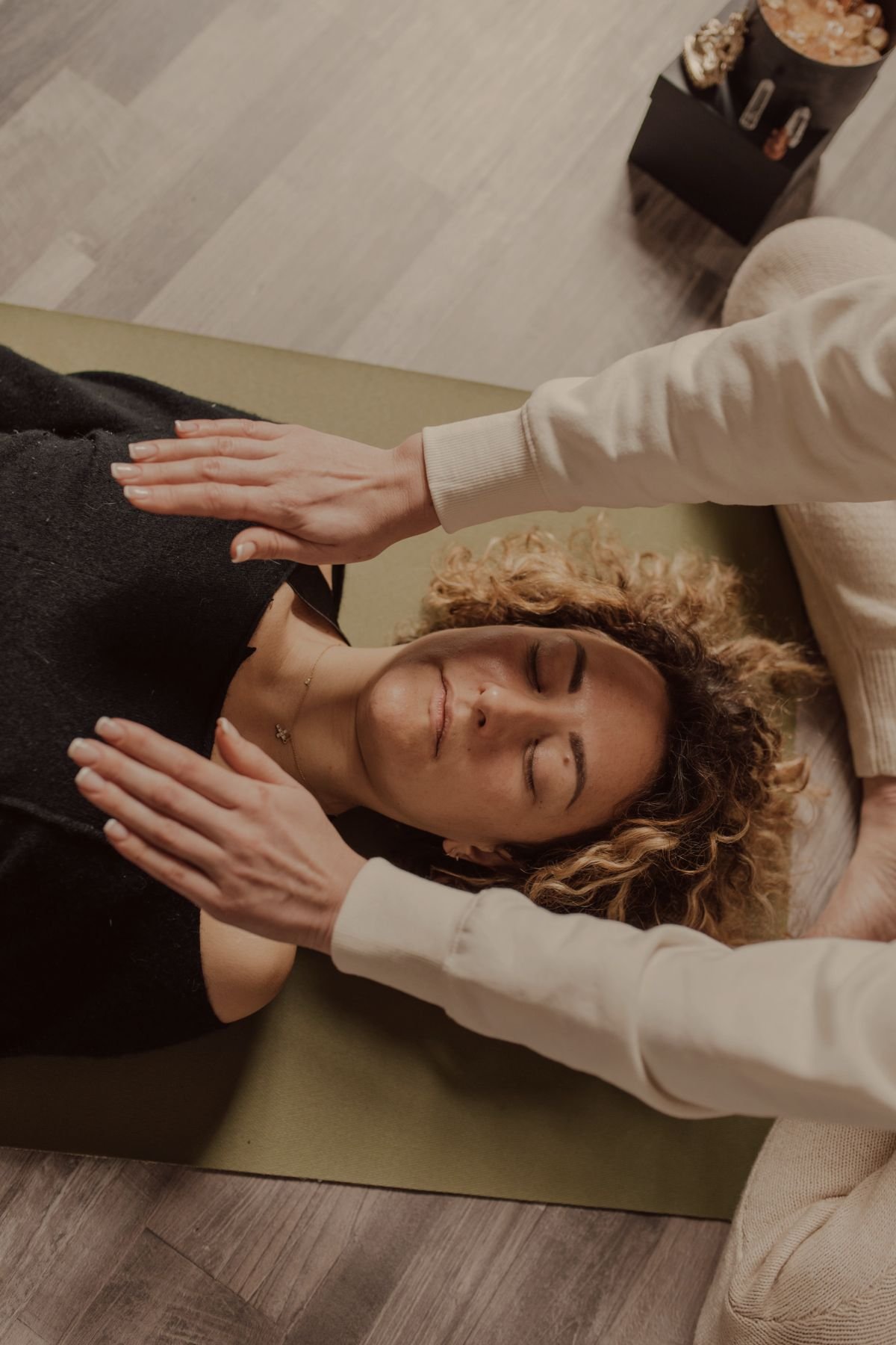 A woman is lying on a mat with her eyes closed, receiving a gentle spinal adjustment from a chiropractor, with her hands resting on her shoulders.