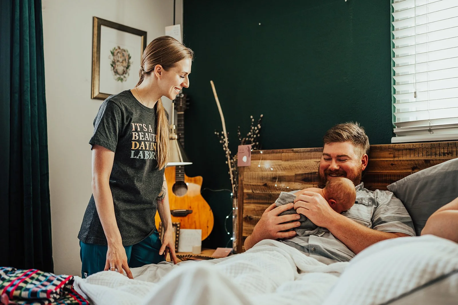 A woman standing next to a man lying in bed, holding a newborn baby. The woman is smiling and looking at the man, who is also smiling. The room has a wooden headboard, a guitar, and decorative lights.