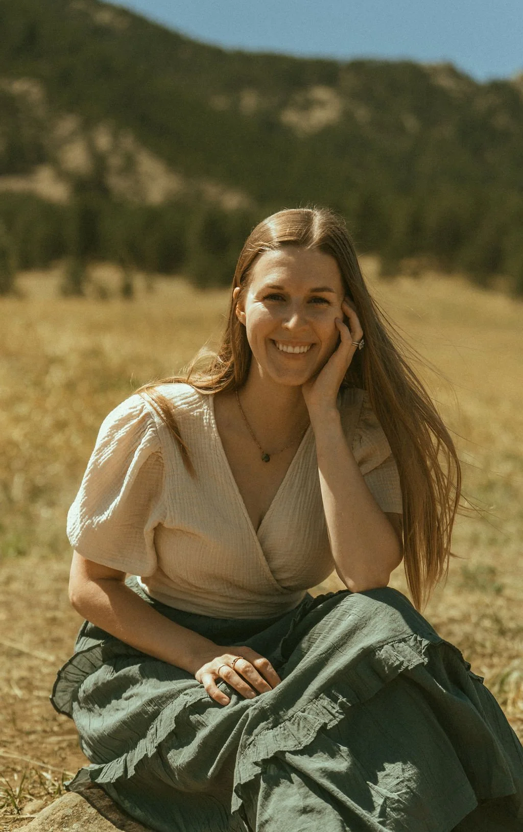 A woman with long brown hair sitting outdoors on a sunny day in a field with mountains in the background. She is smiling, holding her face with one hand, and wearing a light-colored blouse and cargo pants.