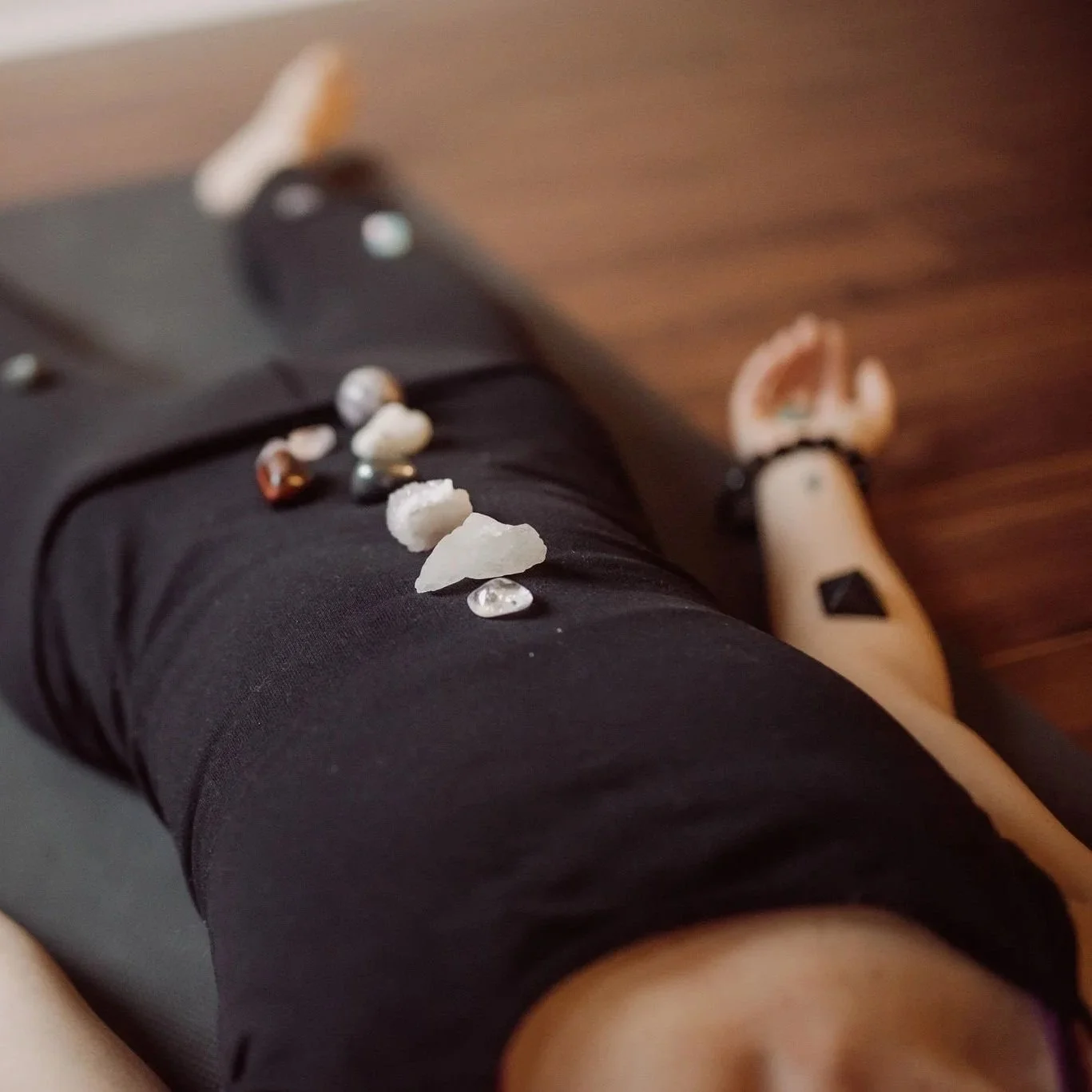 A person's leg wearing black leggings with crystals and stones scattered on it, and a tattoo on the ankle, with a wooden floor in the background.