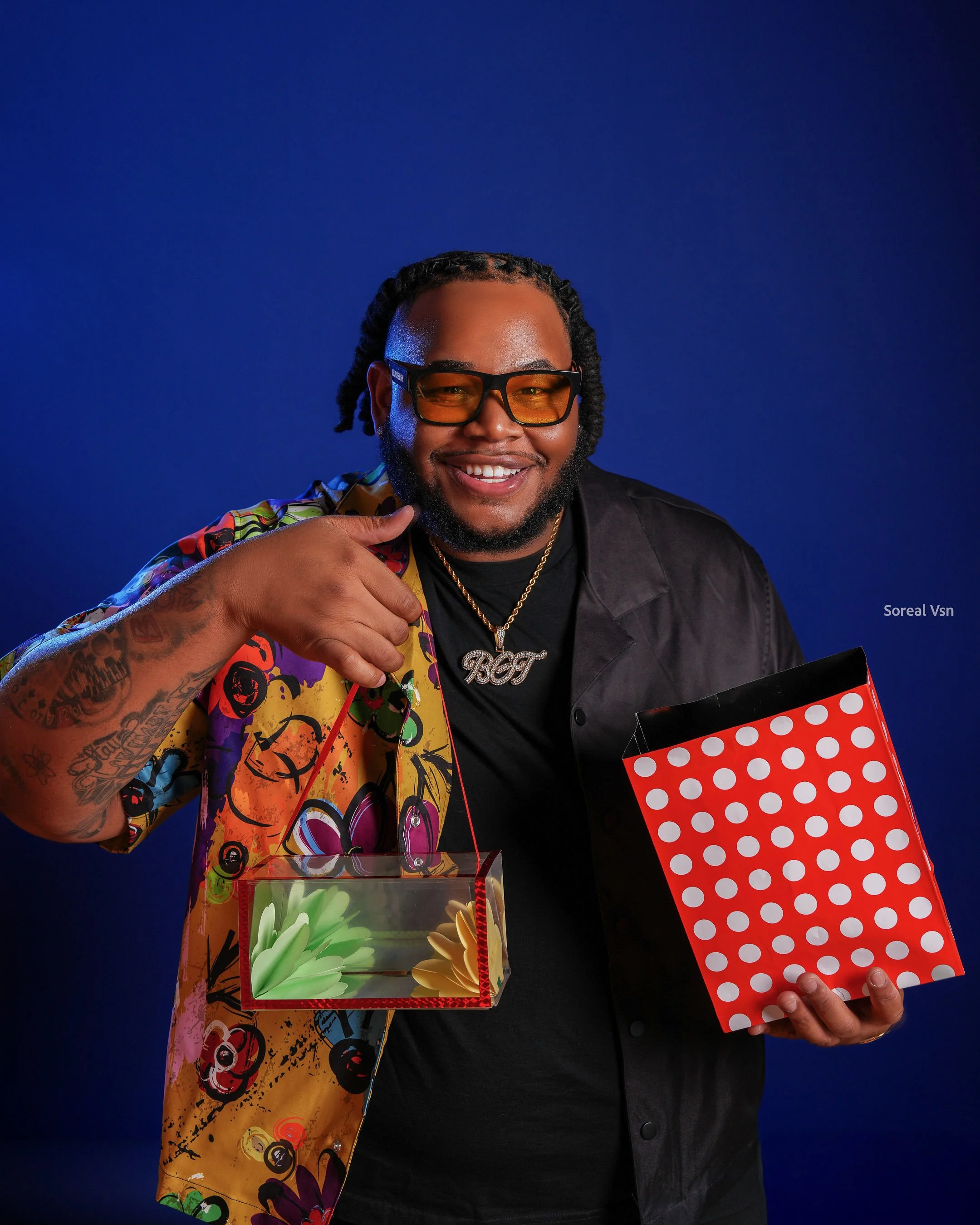 A smiling man with glasses and tattoos holding gift boxes against a blue background.