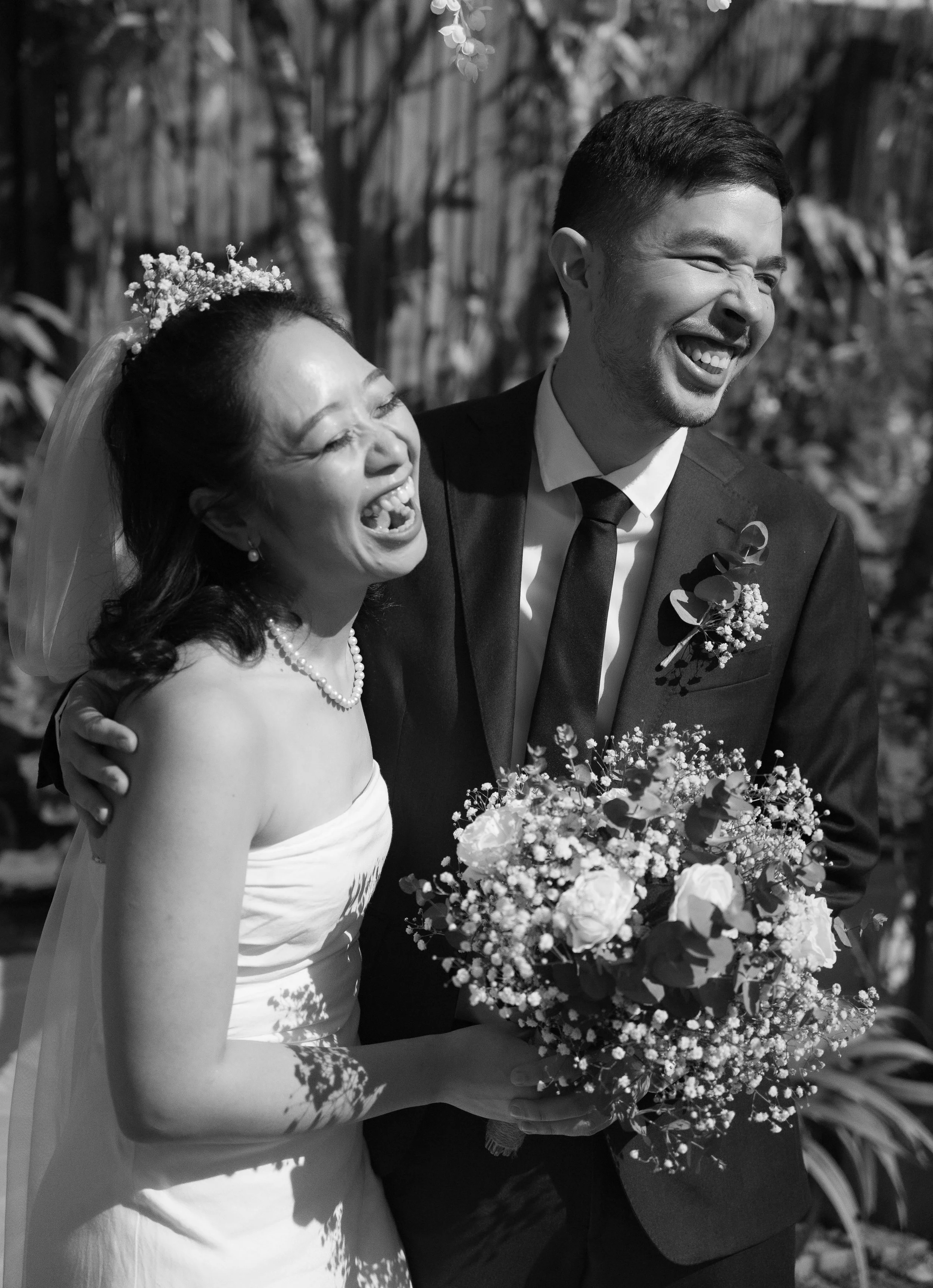Black and white photo of a bride and groom laughing happily, the bride in a strapless dress holding a bouquet of flowers, the groom in a suit with a boutonniere, outdoors with trees and foliage in the background.