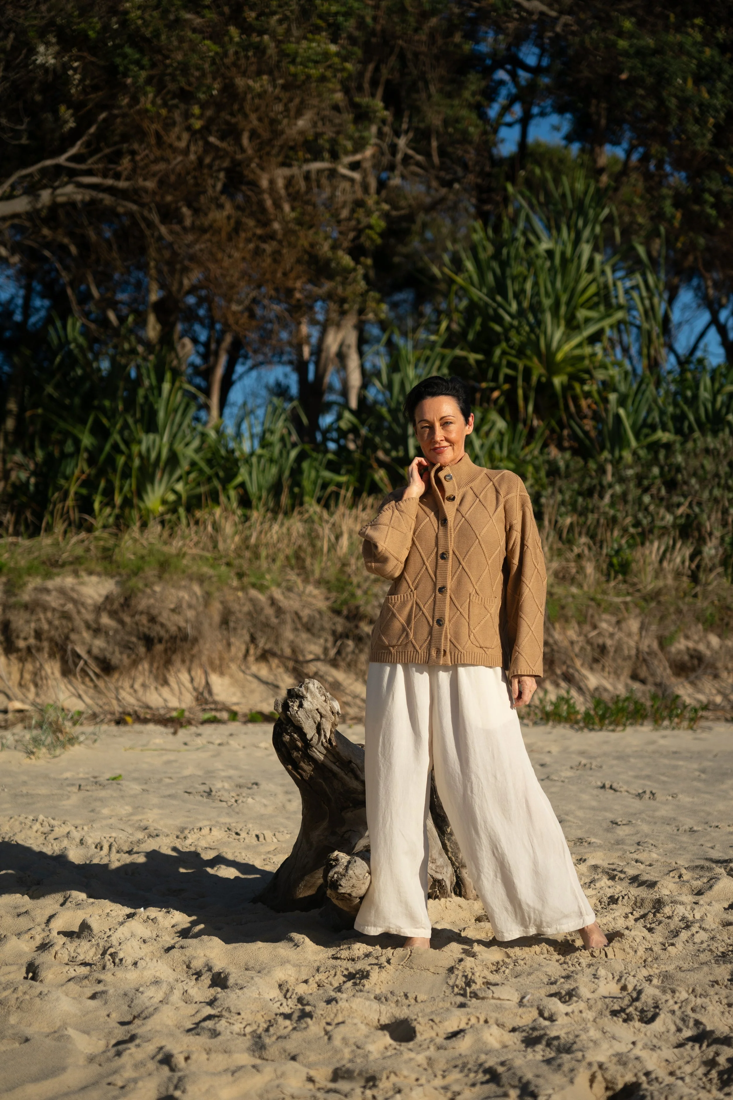 Woman with short dark hair stands on a sandy beach beside a large driftwood stump, wearing a tan diamond-knit button-up cardigan and white wide-leg linen pants, one hand raised near her face, dense coastal bushland behind her.
