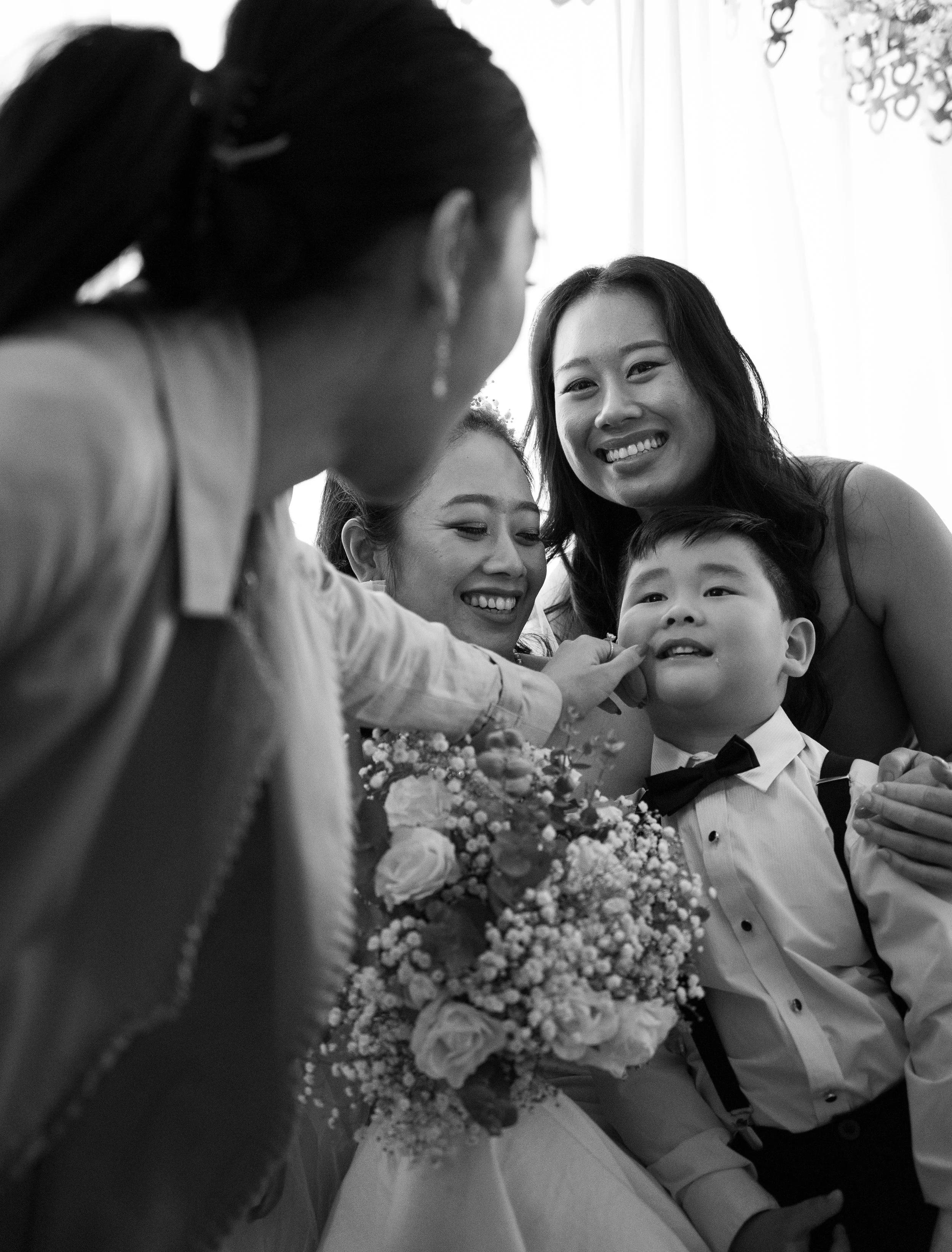 Candid wedding photograph of women and a child sharing a joyful moment indoors, one holding a bouquet of flowers