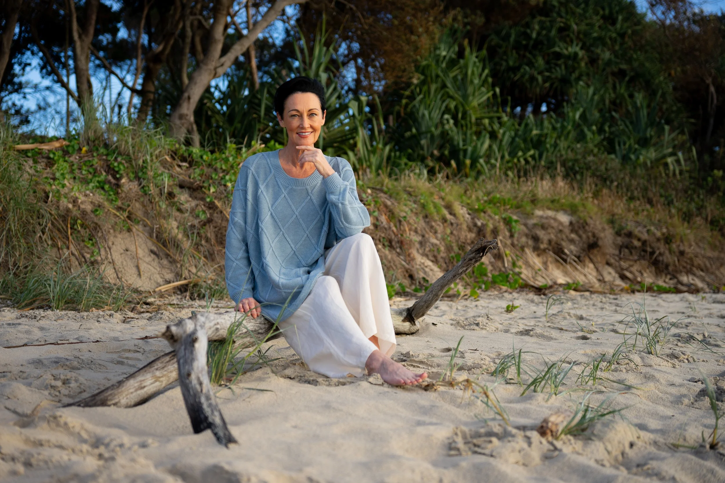 Woman with short dark hair sits on a driftwood log on a sandy beach, wearing an oversized light blue diamond-knit sweater and white wide-leg linen pants, smiling with chin resting on hand, native coastal bushland in background.