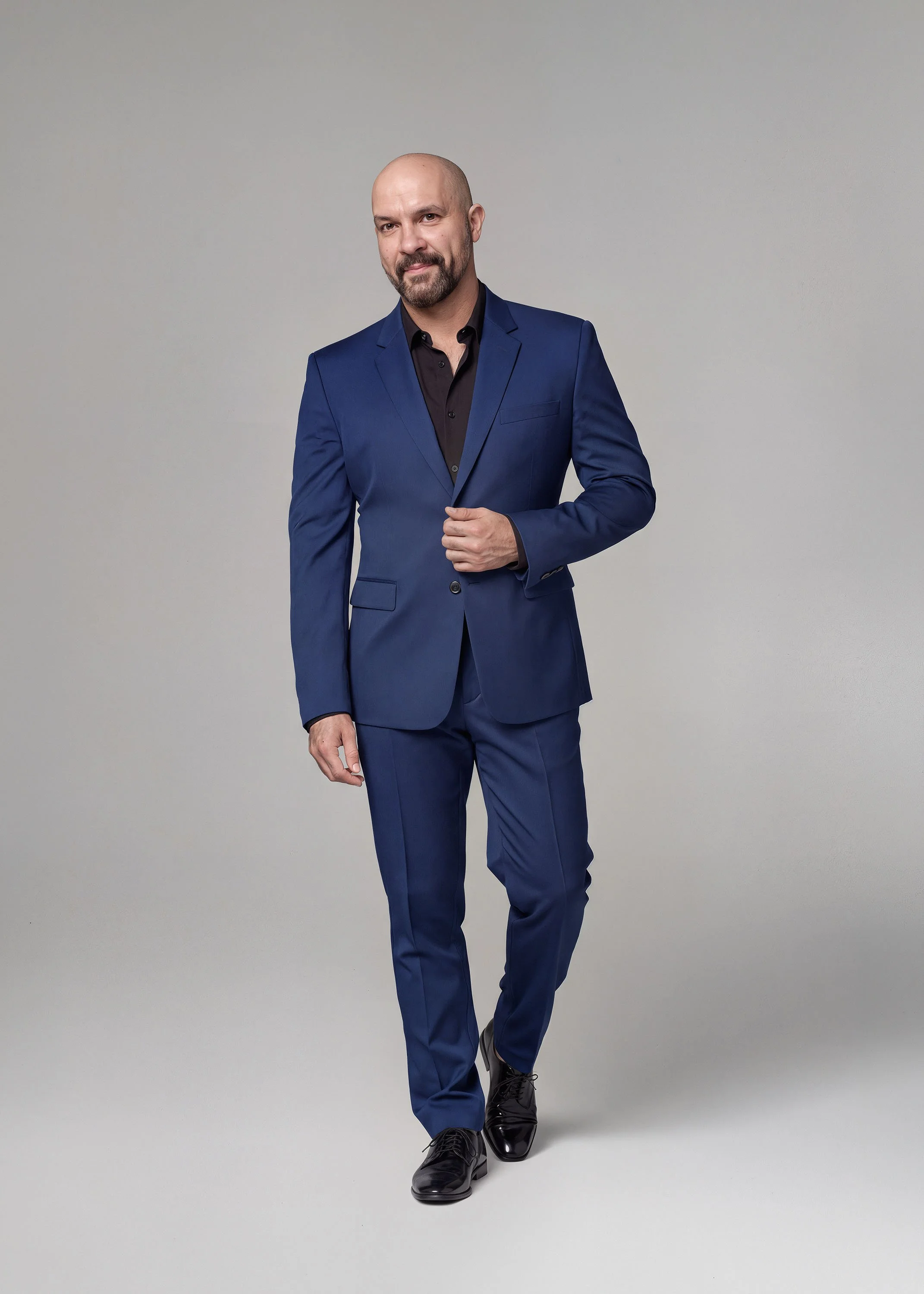 Portrait of Mark Araujo. An elegant man with a beard and shaved head wearing a blue suit, black shirt, and black shoes walking against a plain gray background.