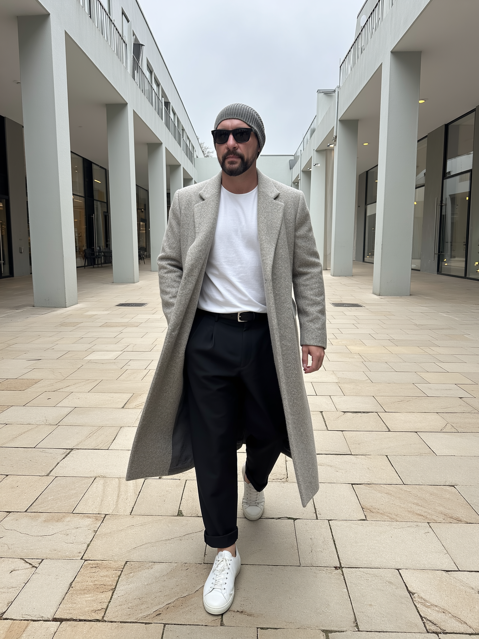 Portrait of Mark Araujo, a man walking outdoors in front of modern white buildings, wearing sunglasses, a gray beanie, a light gray coat, black pants, and white sneakers.