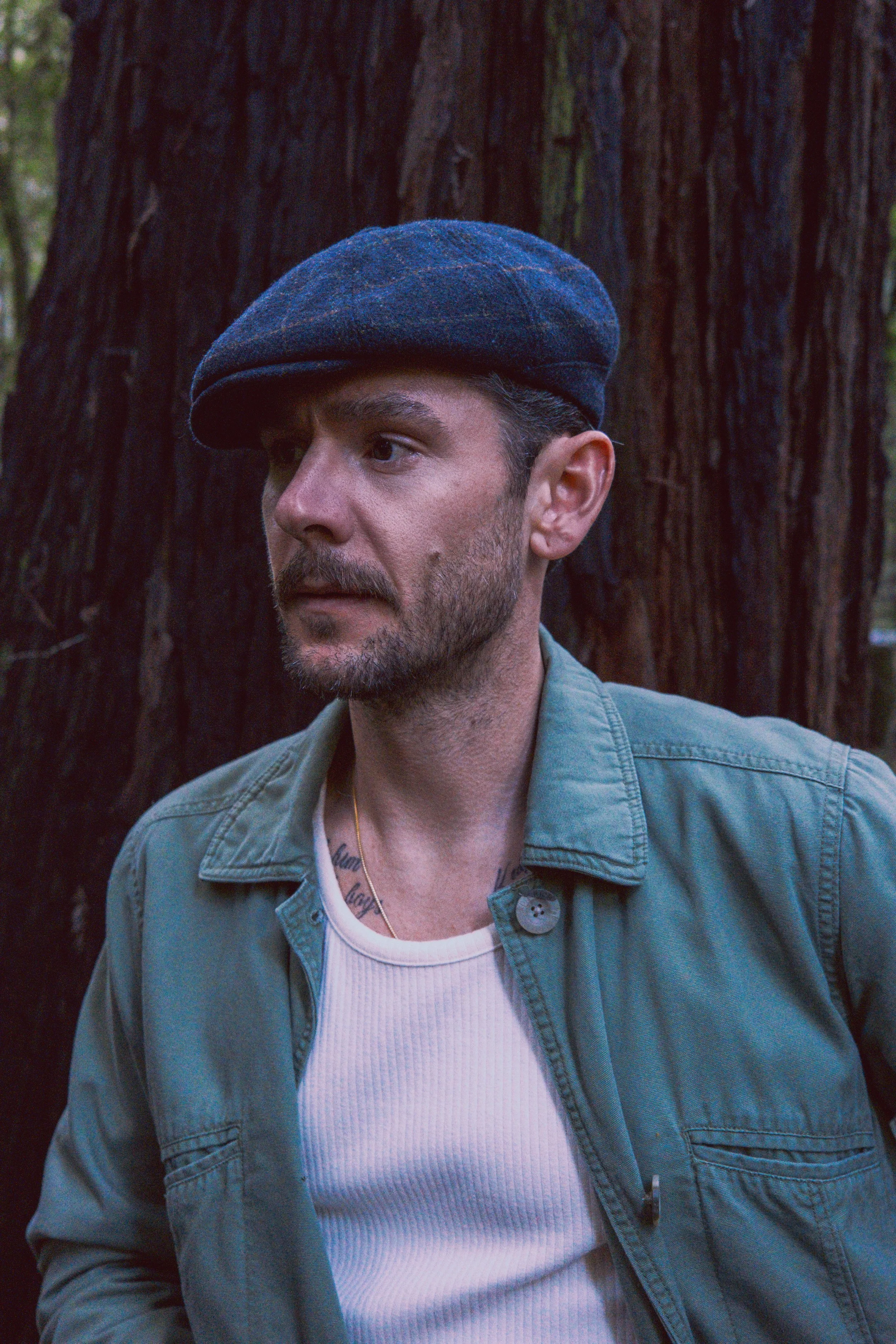 A man wearing a blue flat cap, a light green jacket, and a white tank top, standing outdoors in front of a large tree with textured bark.