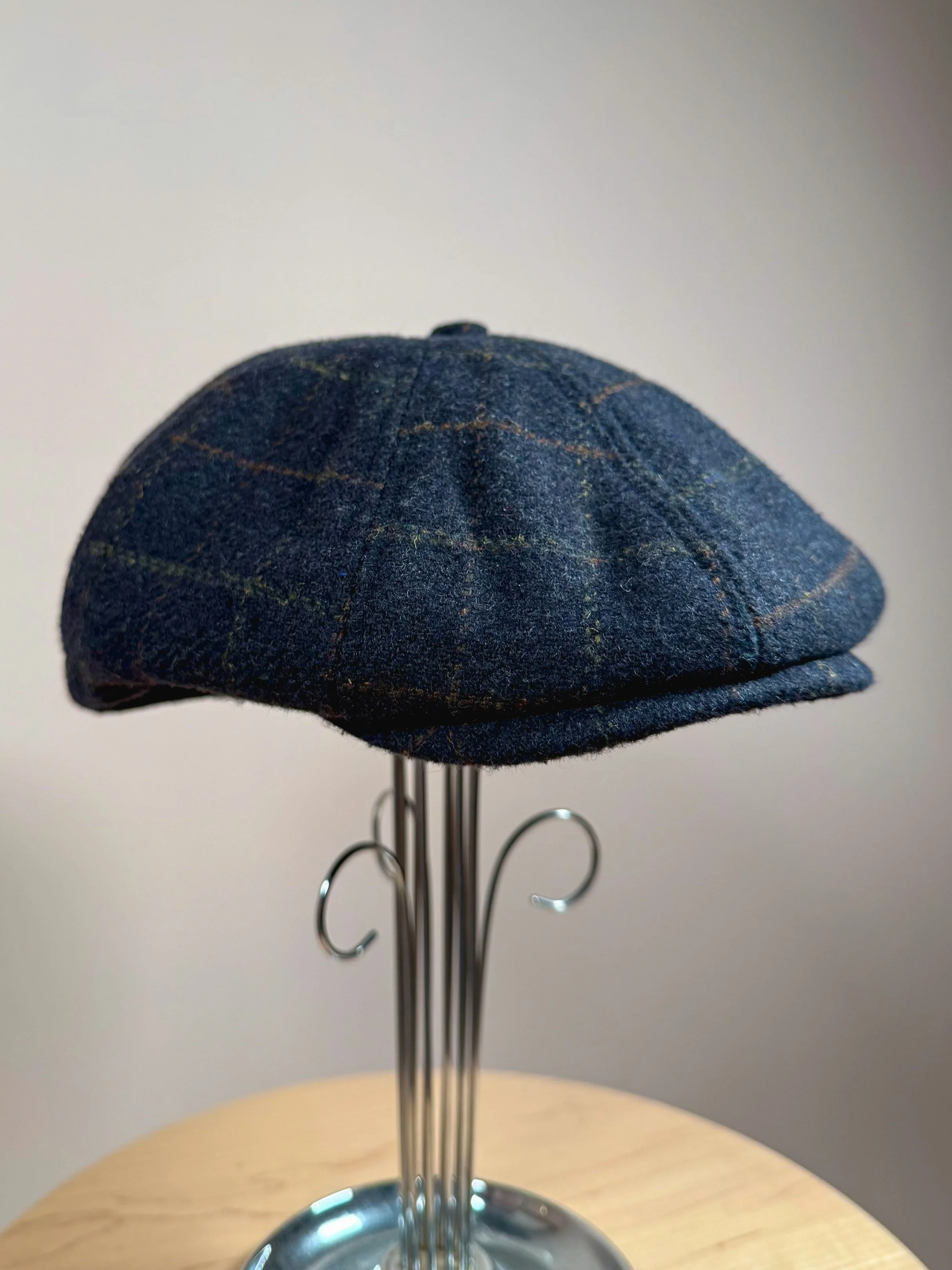 A blue plaid flat cap displayed on a metal stand with a wooden surface and plain gray background.
