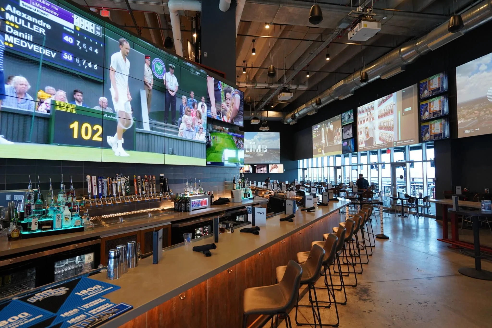 Interior of a sports bar with a long counter, multiple large TV screens showing a tennis match, empty chairs, and a variety of beverages behind the bar.