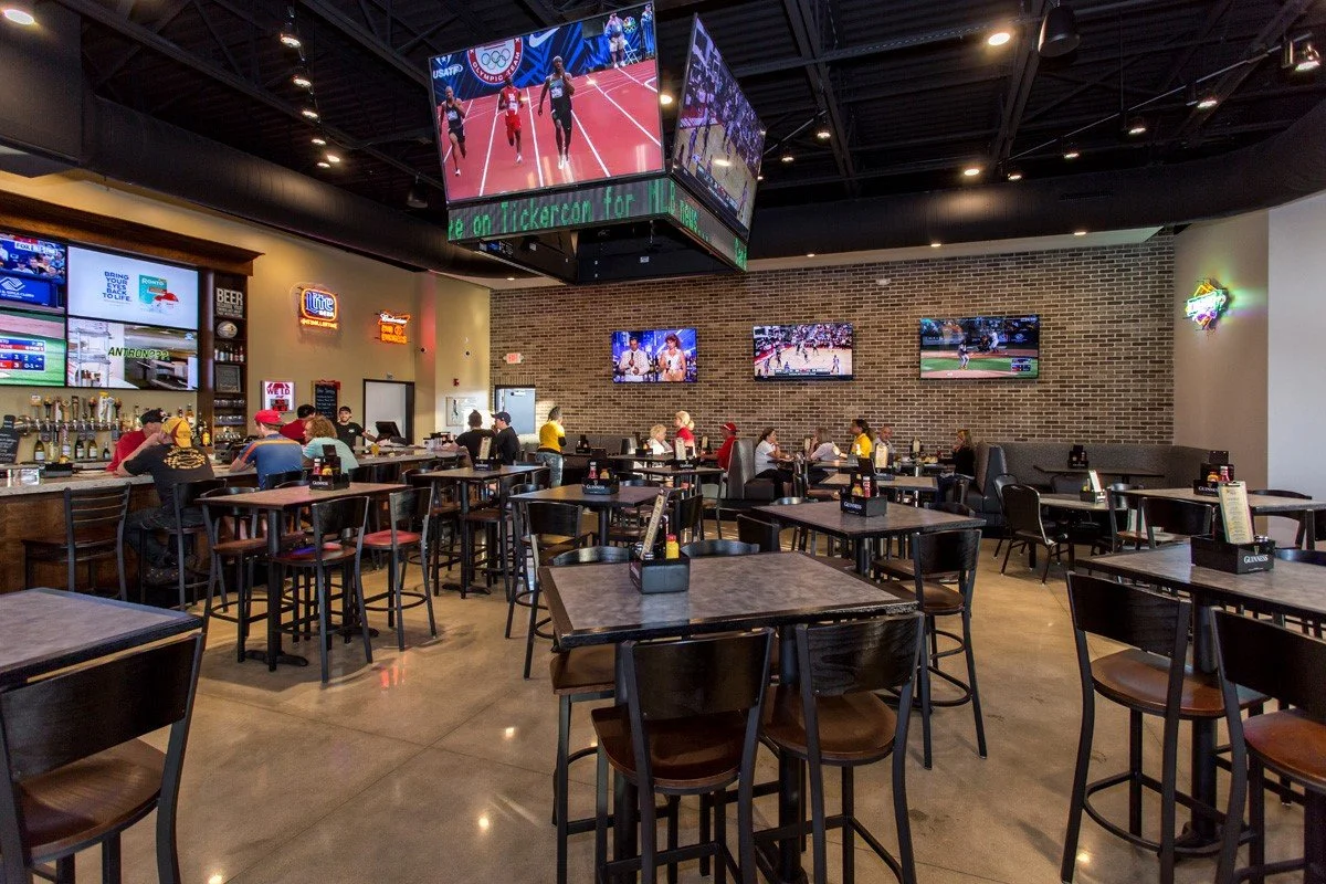 A sports bar with multiple tables and chairs, several TVs on the brick wall showing various sports games, and a bar area with taps on the left side. There are people sitting and watching the screens.