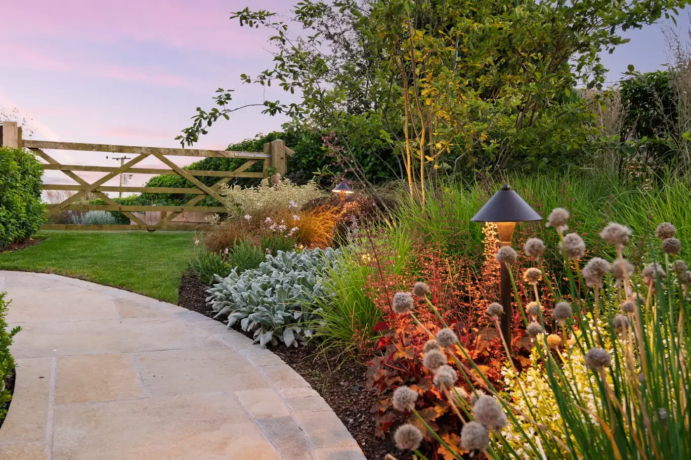 A cozy backyard with outdoor furniture, a small fire pit, surrounded by lush greenery and illuminated garden lights at dusk.