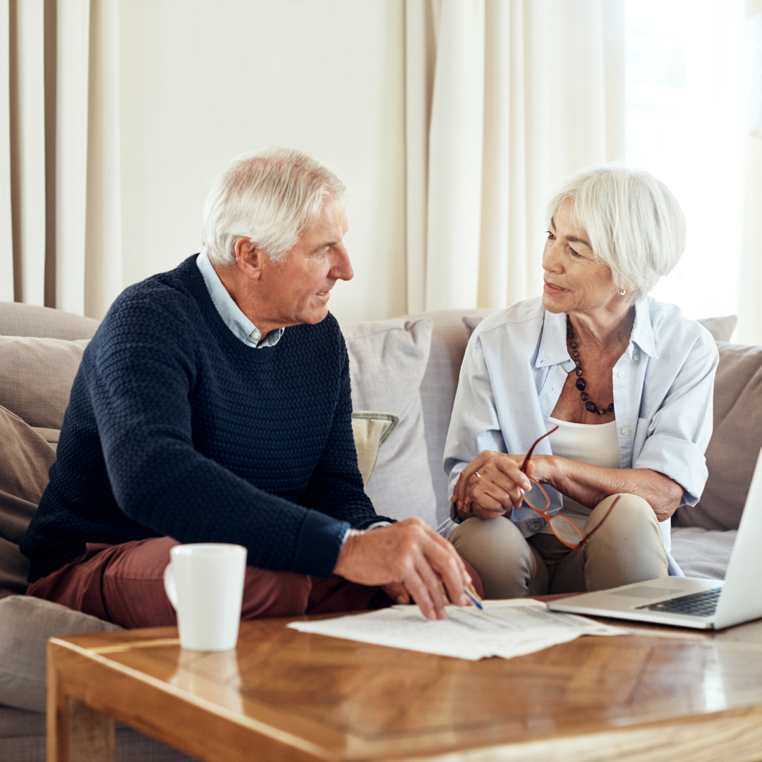 Older man and woman sitting on a couch having a discussion with papers, a laptop, and a coffee mug on the table in front of them.