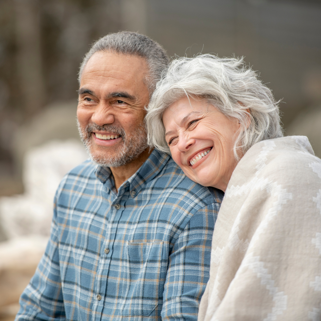 An elderly couple smiling and cuddling outdoors on a cloudy day.