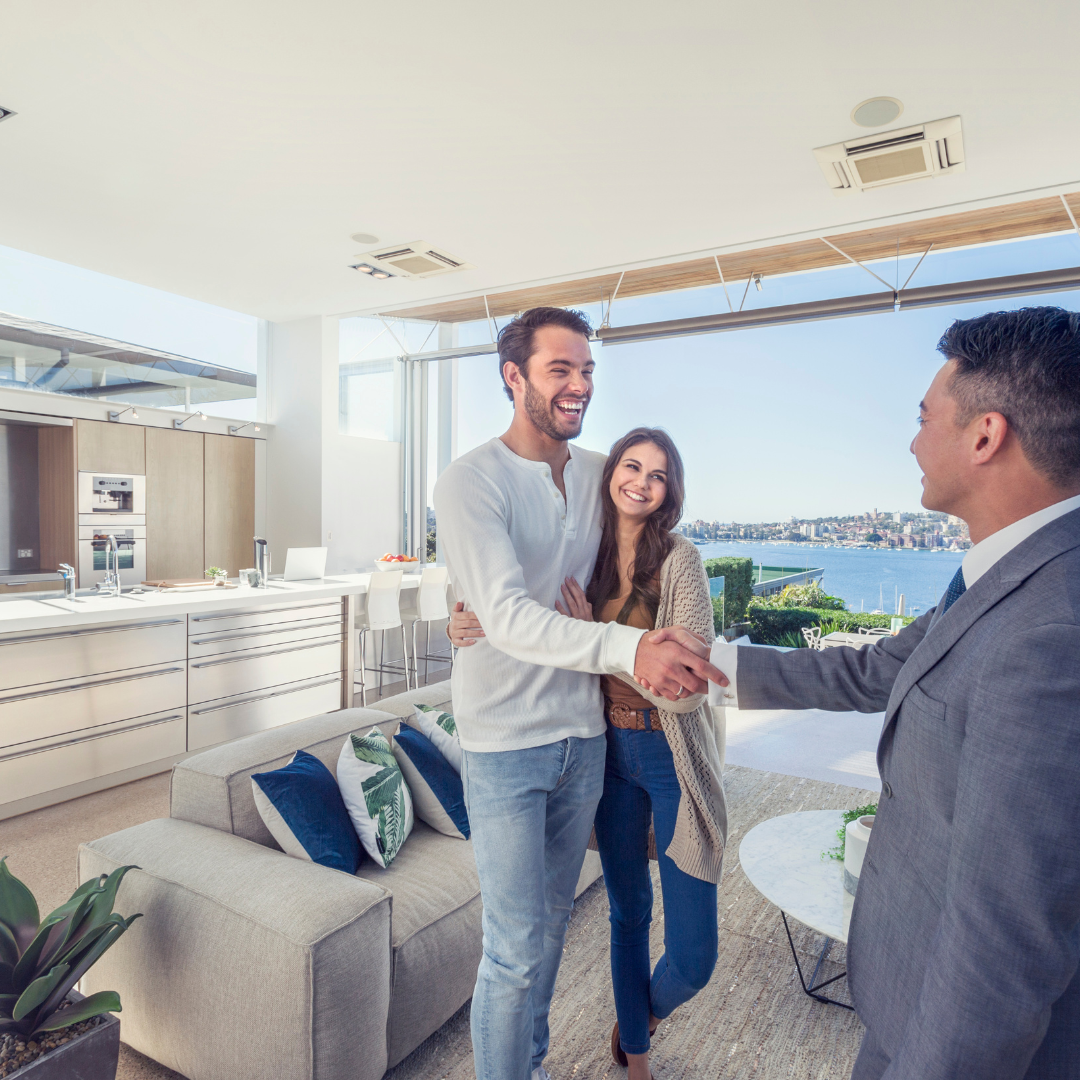 Two men shaking hands and smiling, with a woman standing behind them, in a bright modern living room with a view of water and city.