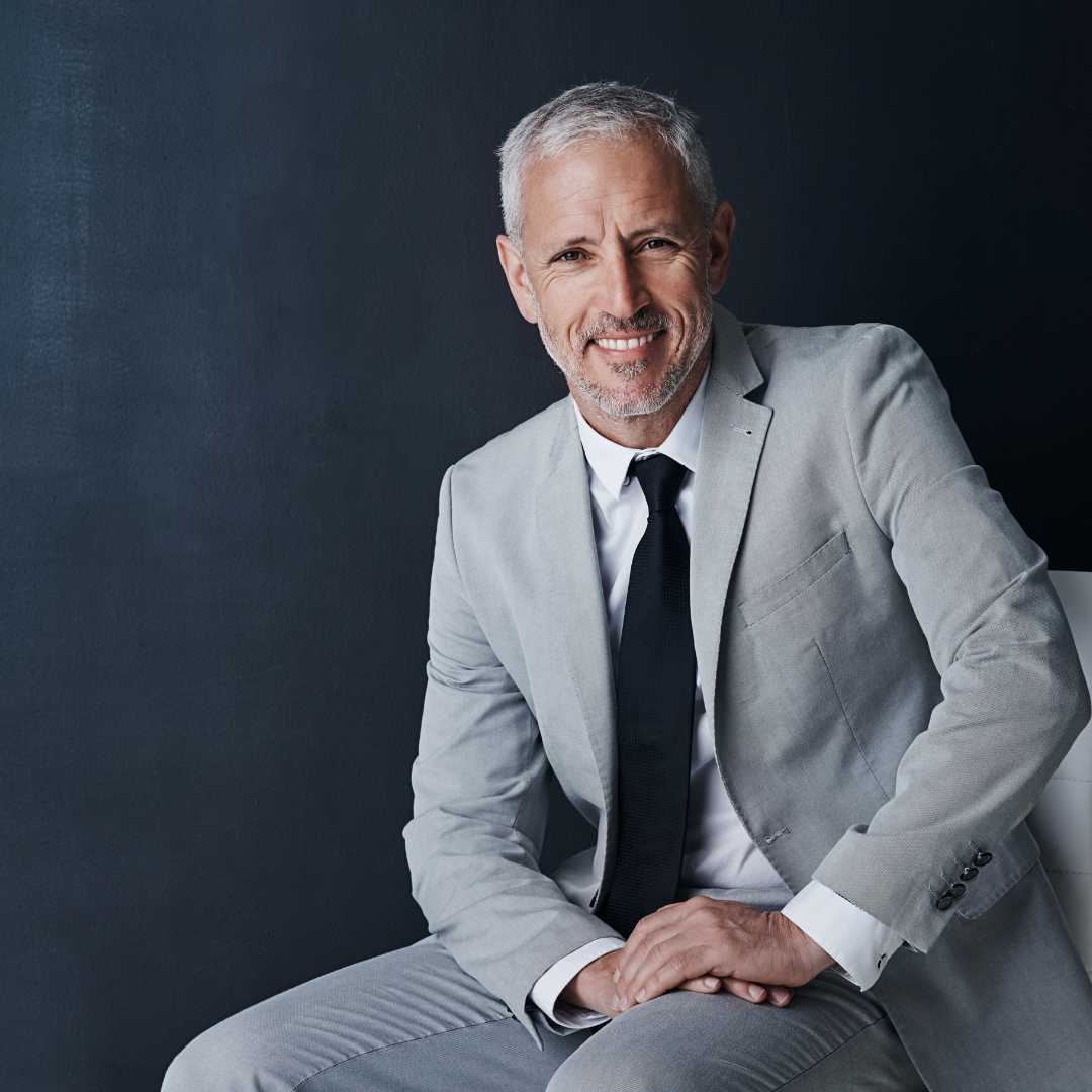 A smiling middle-aged man with gray hair and beard, wearing a light gray suit, white shirt, and black tie, sitting against a dark wall.