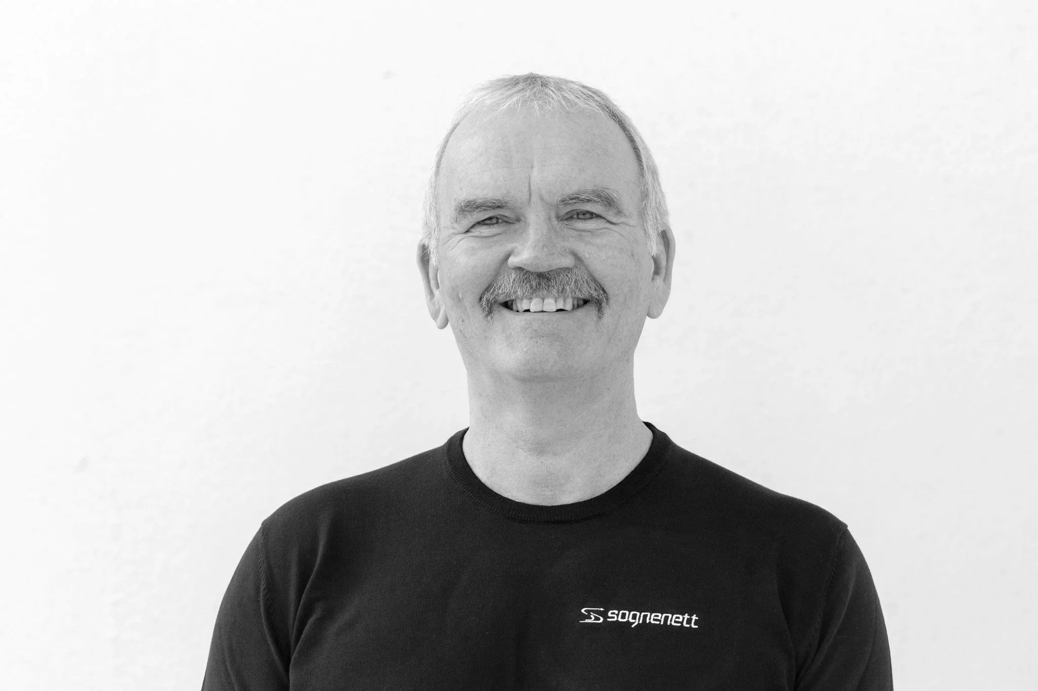 Portrett av en mann med blankt hår, bart og smil, iført en mørk skjorte, mot en mørk bakgrunn.