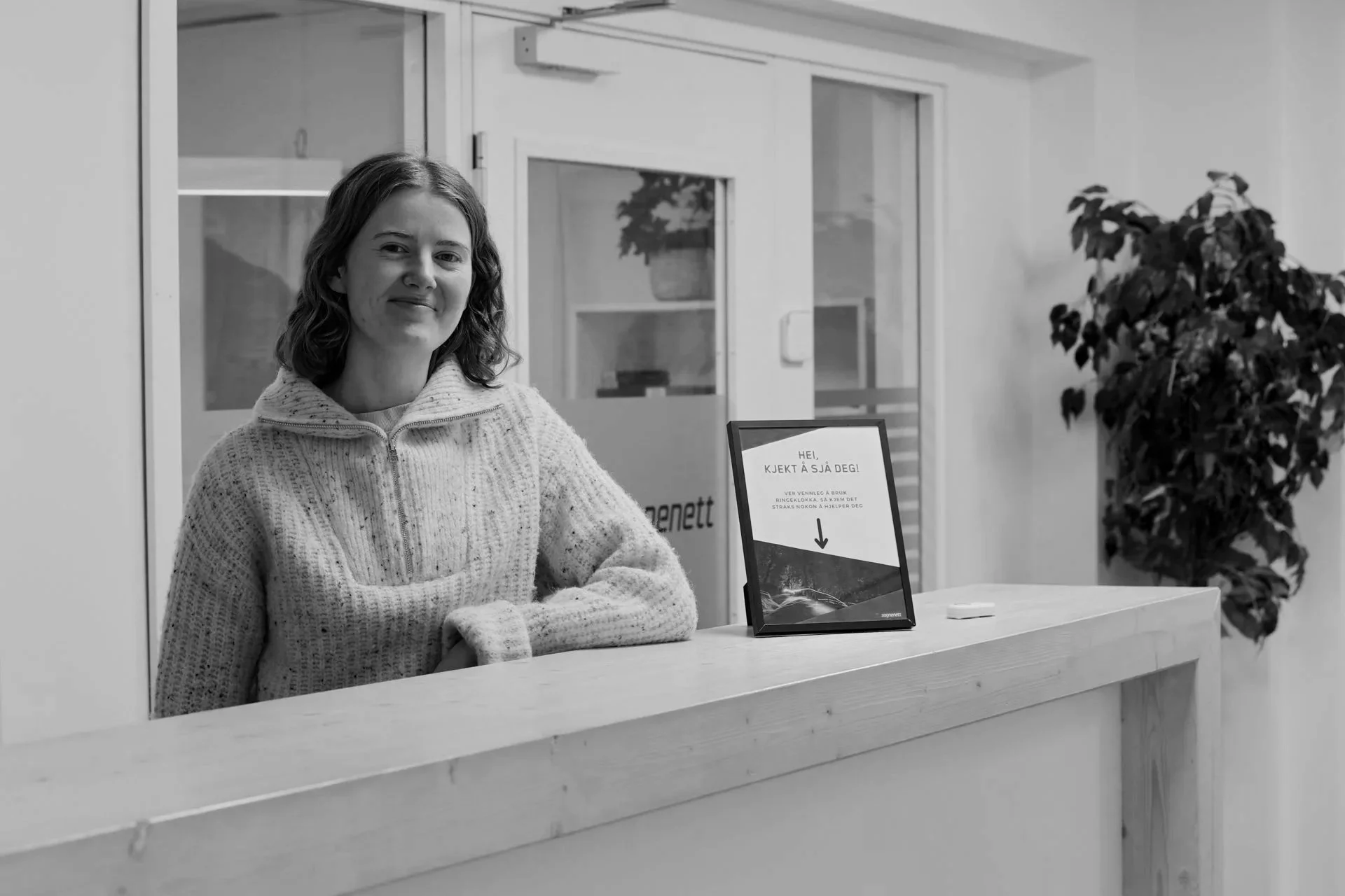 En kvinne smiler mens hun sitter bak en resepsjonsdisk, foran en digital skjerm med informasjon, i et rom med hvite vegger og et stort plante.