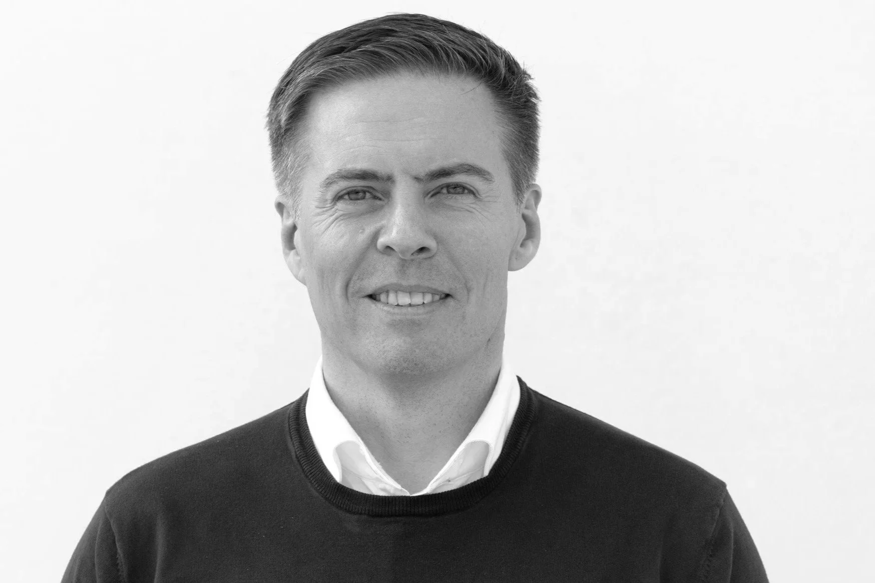 Et svart-hvitt portrett av en mann med briller og helstøpt hår, iført svart skjorte, mot en lys trevegg.