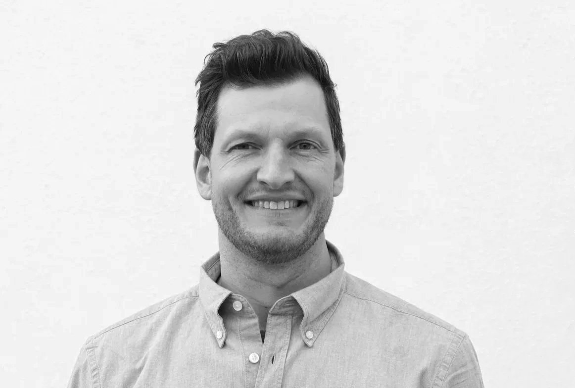 Svart-hvitt fotografi av en smilende mann med kort, mørkt hår, kledd i hvit polo.