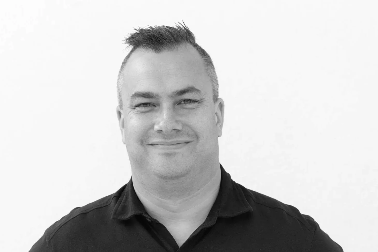 Svart-hvitt fotografi av en smilende mann med kort, mørkt hår, kledd i hvit polo.