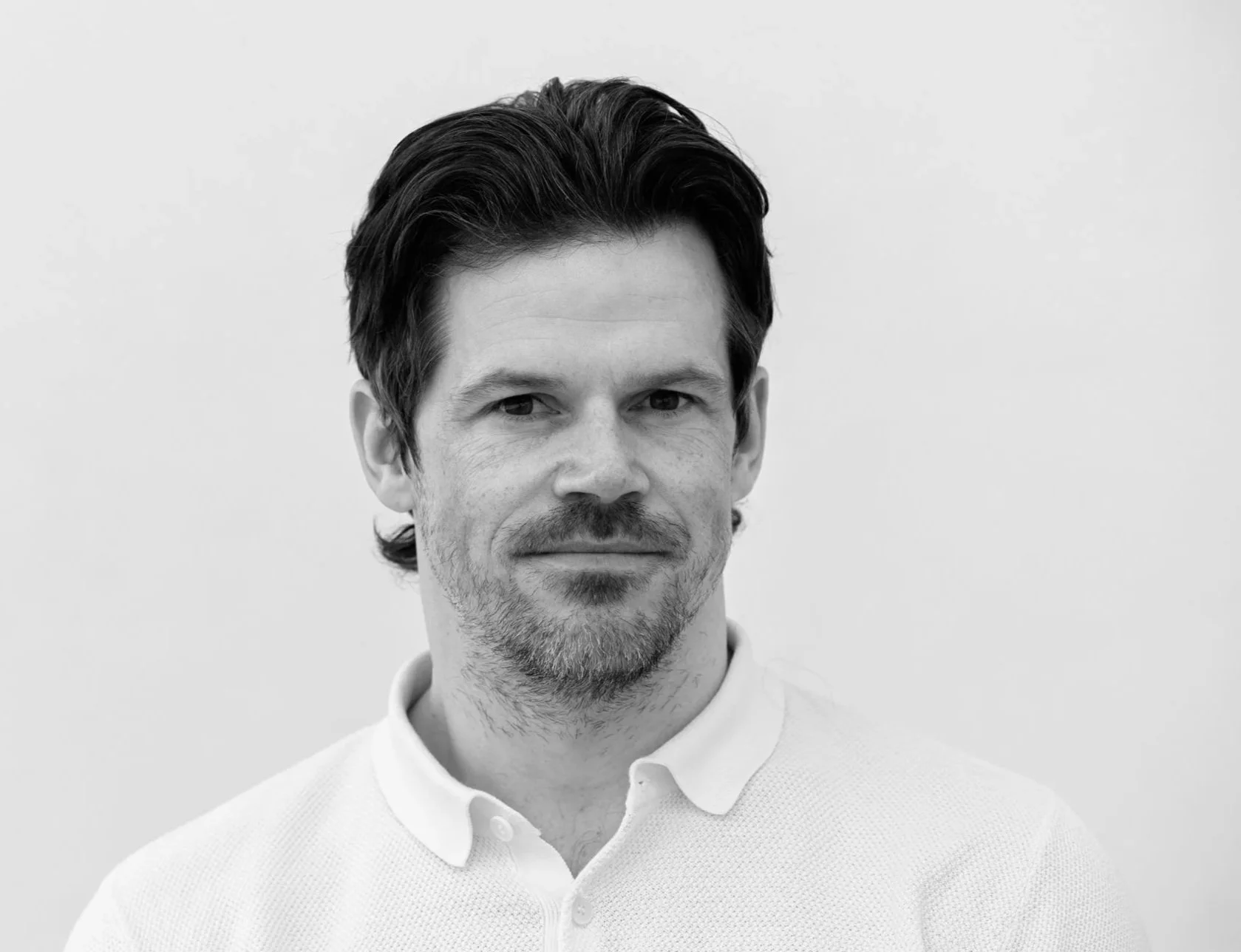 Svart-hvitt bilde av en smilende mann med kort, mørkt hår, iført en hvit polo-skjorte.