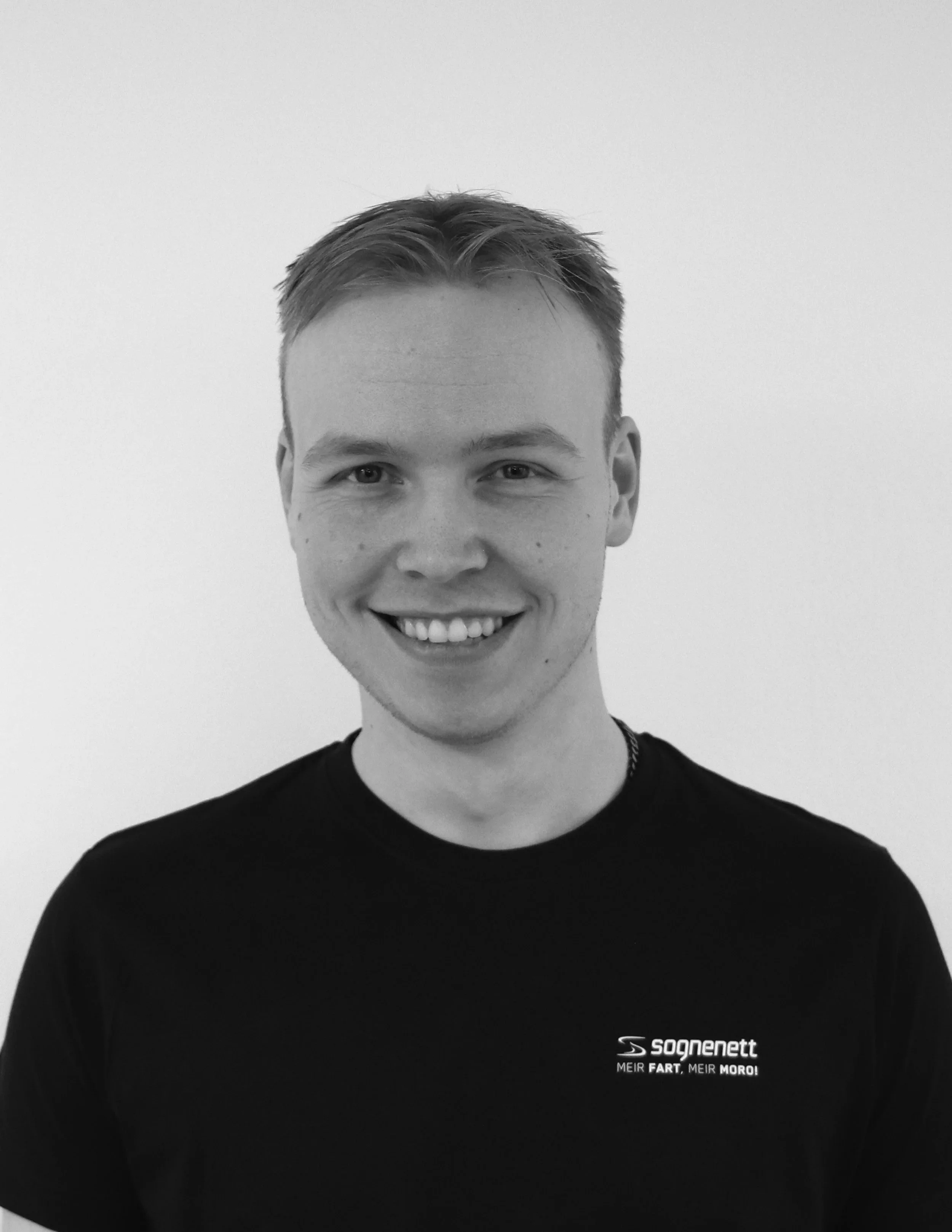 Svart-hvitt bilde av en smilende ung mann med kort hår, iført en svart t-skjorte med logo og tekst.