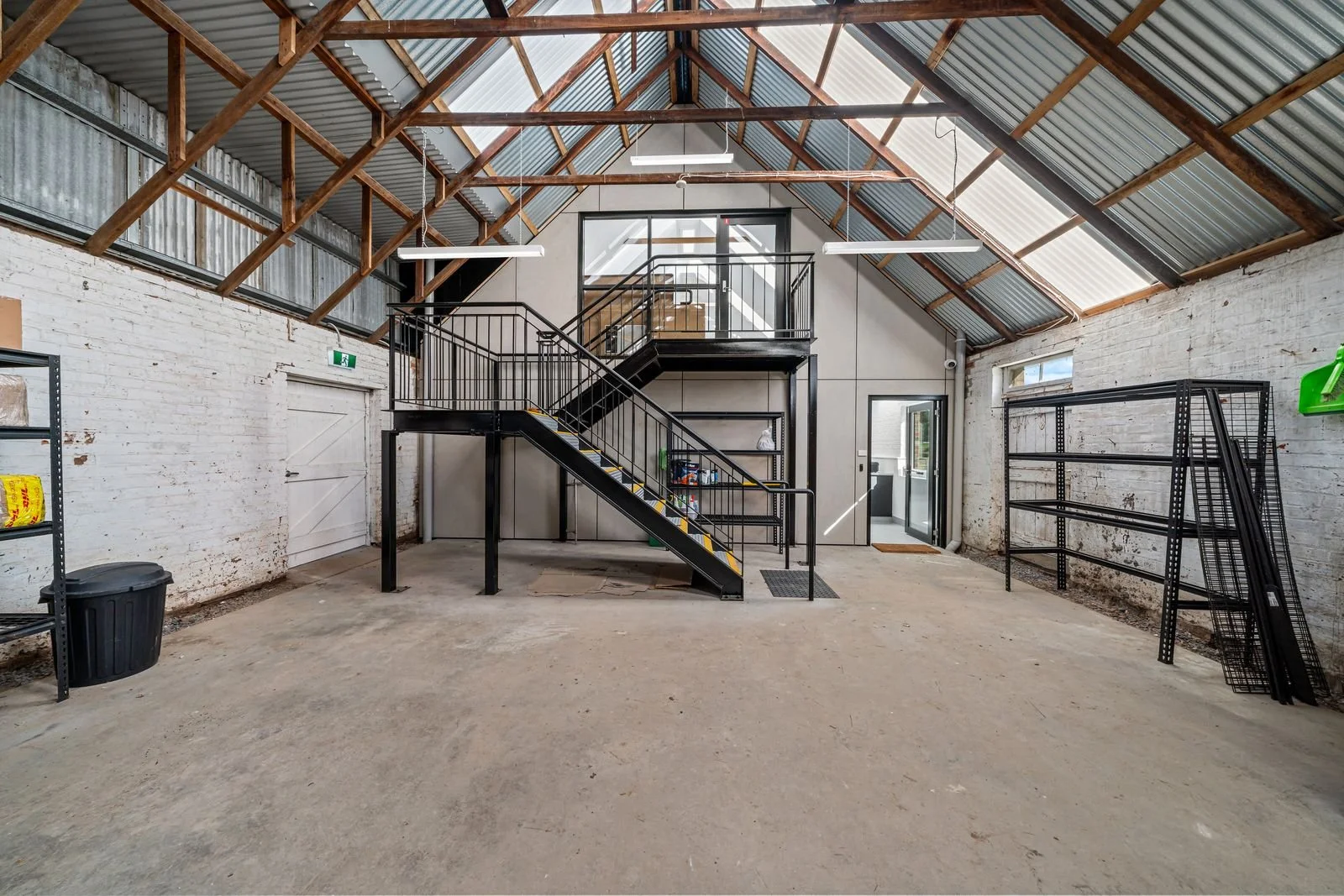 An industrial-style, partially finished garage or workshop with a metal staircase leading to a small second-floor balcony, exposed brick walls painted white, a concrete floor, and a high ceiling with wood and metal beams.