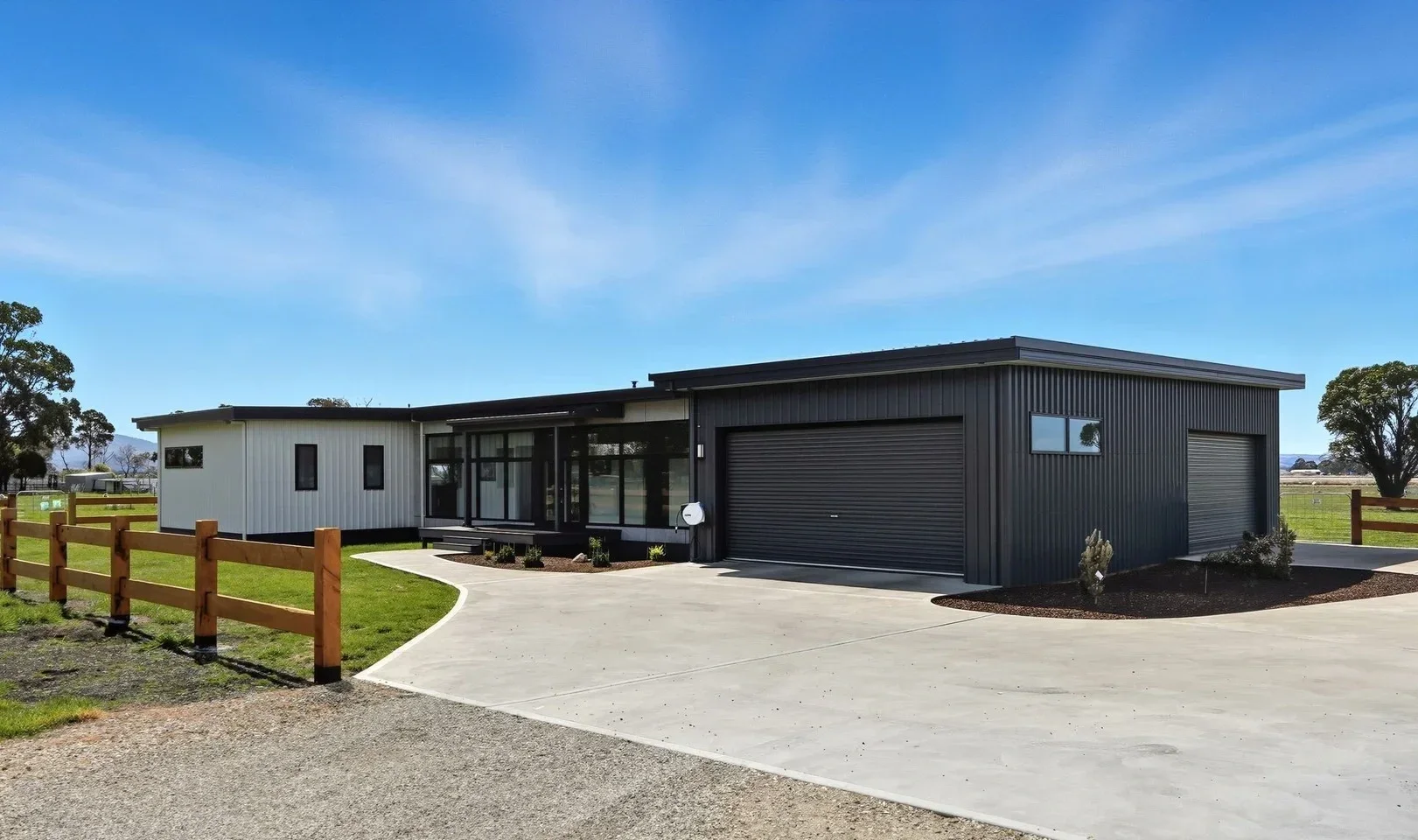 Modern house with black and white exterior, large glass windows, a driveway, and a flagpole in a rural setting under a clear blue sky.