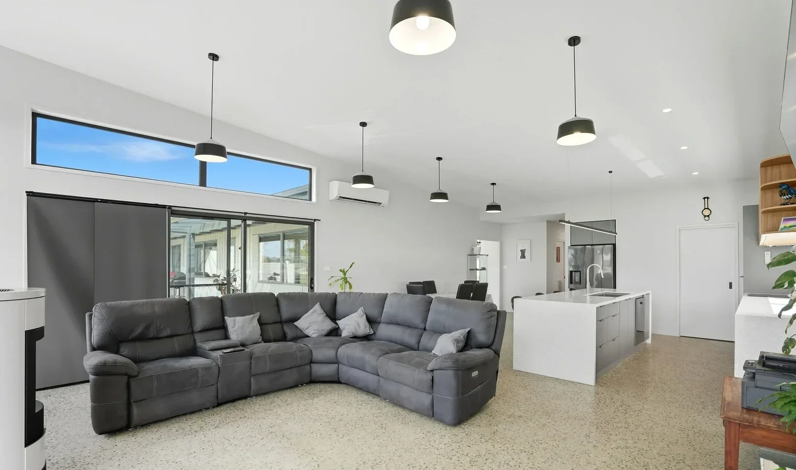 Modern living room with black sectional sofa, white cushions, and high ceiling with pendant lights, open kitchen with island, sliding glass door, and large window.