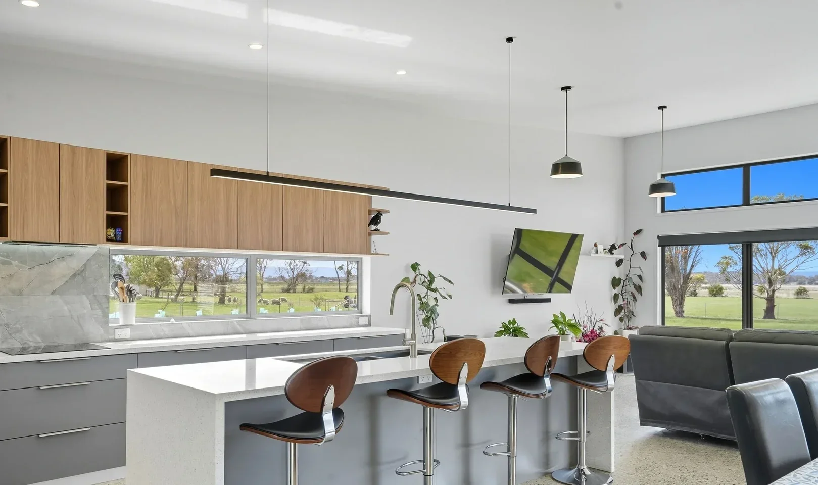 Modern open-concept kitchen and living area with large windows showing a green landscape, bar stools at a kitchen island, flat-screen TV on the wall, and contemporary decor.