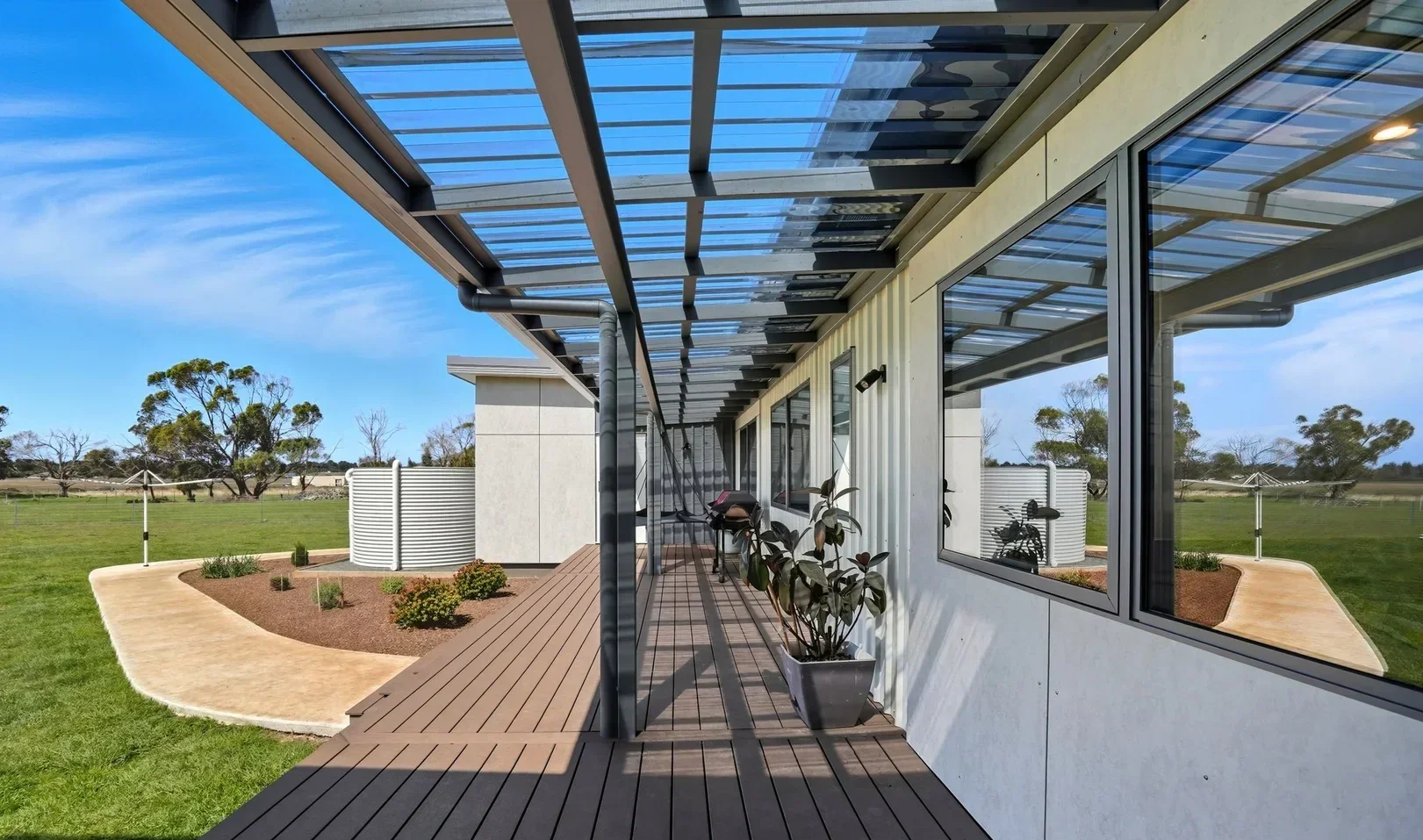 View of a modern house patio with a wooden deck, large windows, potted plants, and a metal pergola with a transparent roof, overlooking a green field with trees.