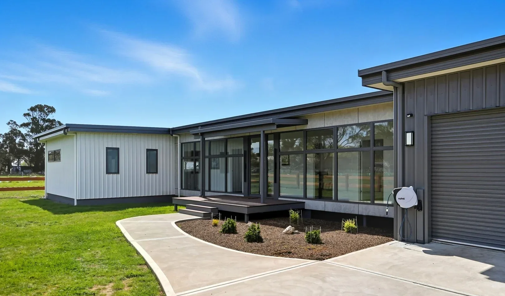 Modern house with gray and white exterior, large glass windows, small porch, landscaped yard, and blue sky.