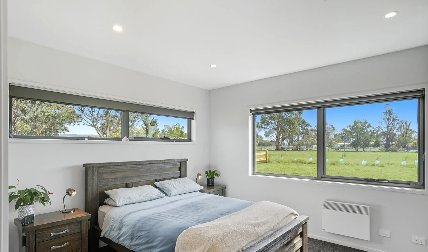A bedroom with a wooden bed frame, white bedding, and two pillows, flanked by two nightstands with lamps and plants, and large windows showing green trees and a blue sky outside.