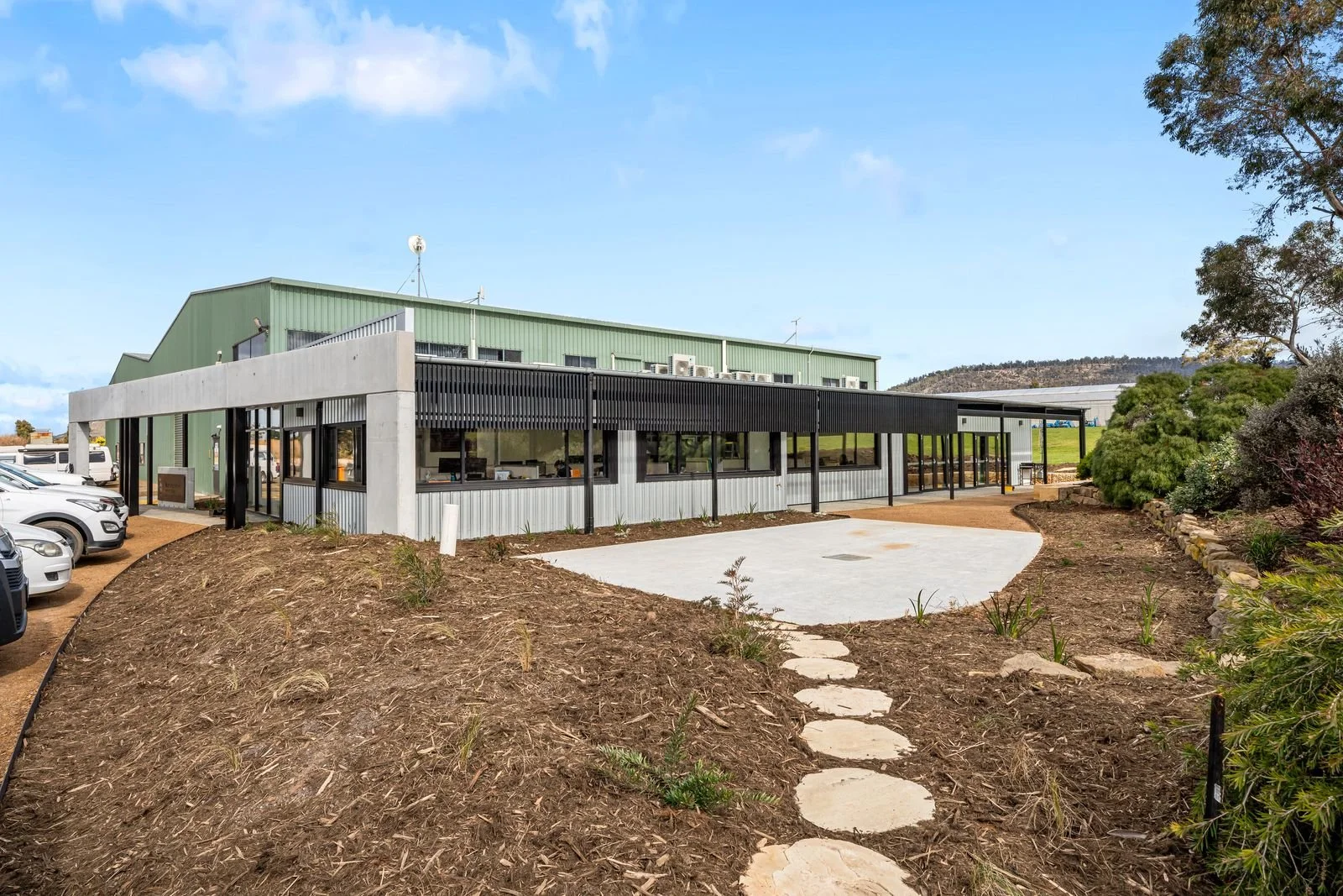 Modern building with large glass windows and a green metal roof, parking lot with white cars, landscaped area with a stone pathway, trees, and a hillside in the background.