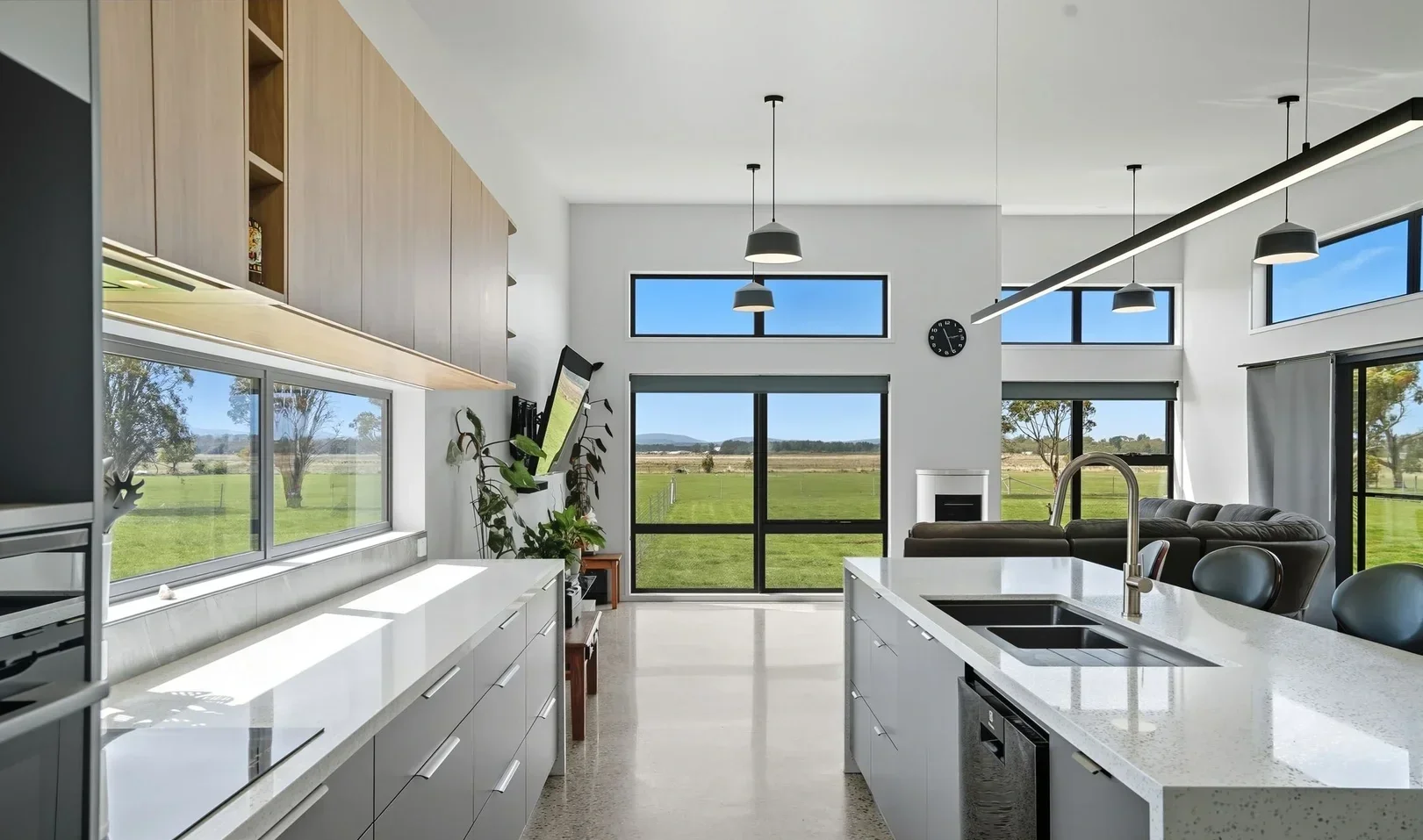 Modern kitchen with large windows, white countertops, and light wood cabinets, overlooking a green outdoor field.