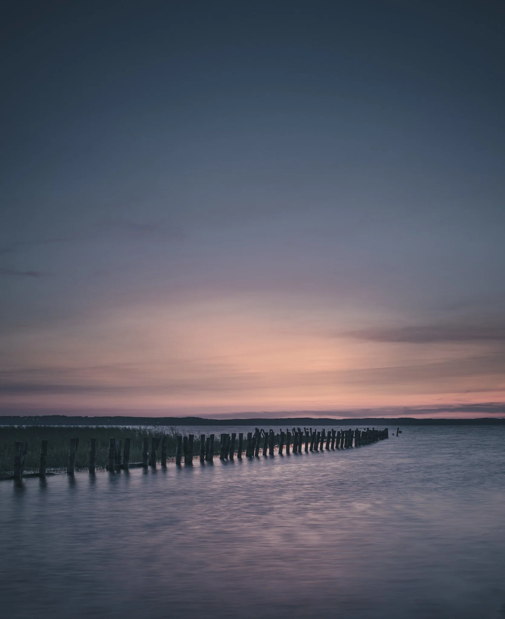 A calm body of water at sunset with a line of wooden posts extending into the distance against a colorful sky.
