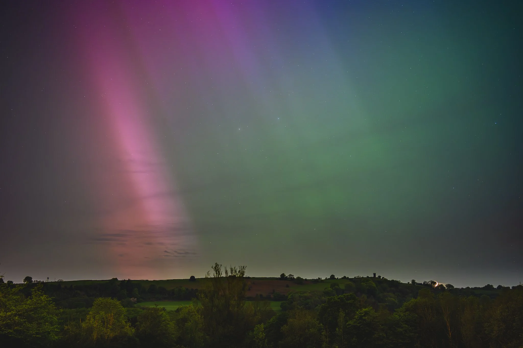 Night sky with colorful aurora borealis over a green landscape with trees and hills
