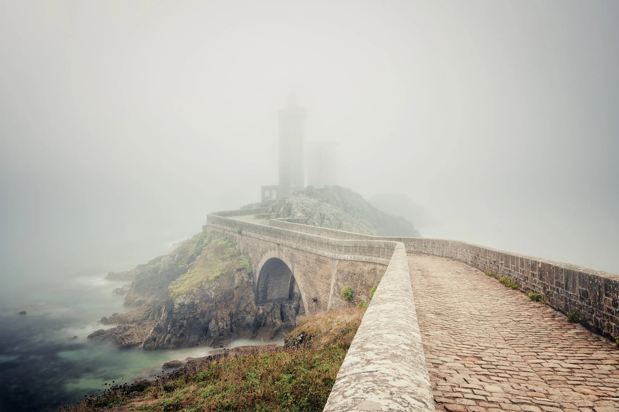 A foggy coastal pathway with a brick pedestrian walkway and stone railing leading to a lighthouse on a rocky cliff.