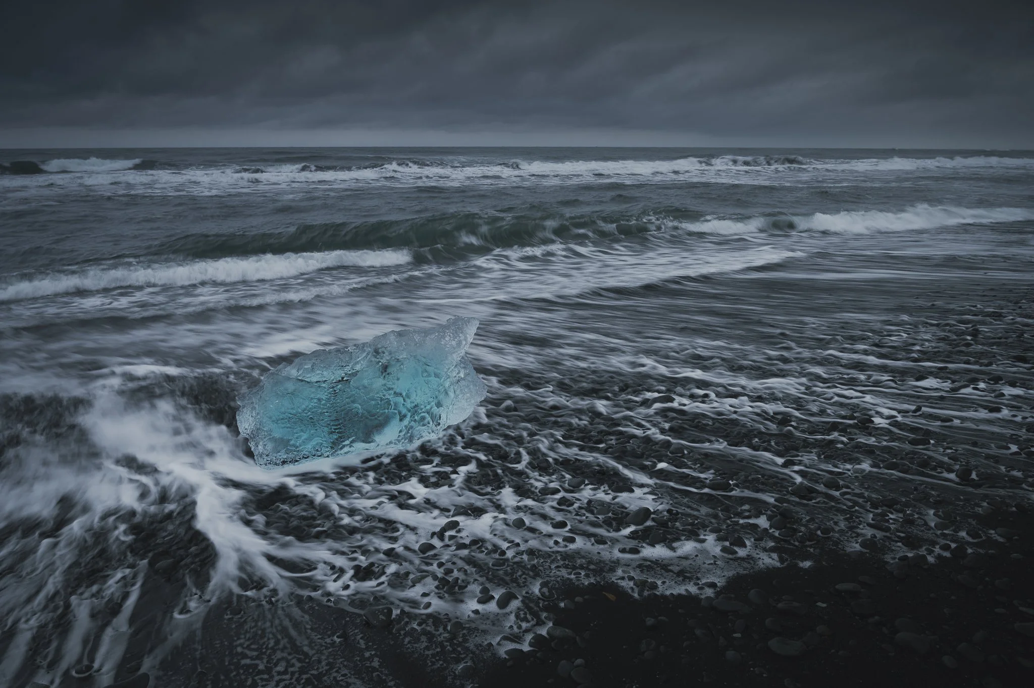 Dark stormy sky over an ocean with waves crashing on a black pebble beach, with a blue iceberg partially submerged near the shoreline.