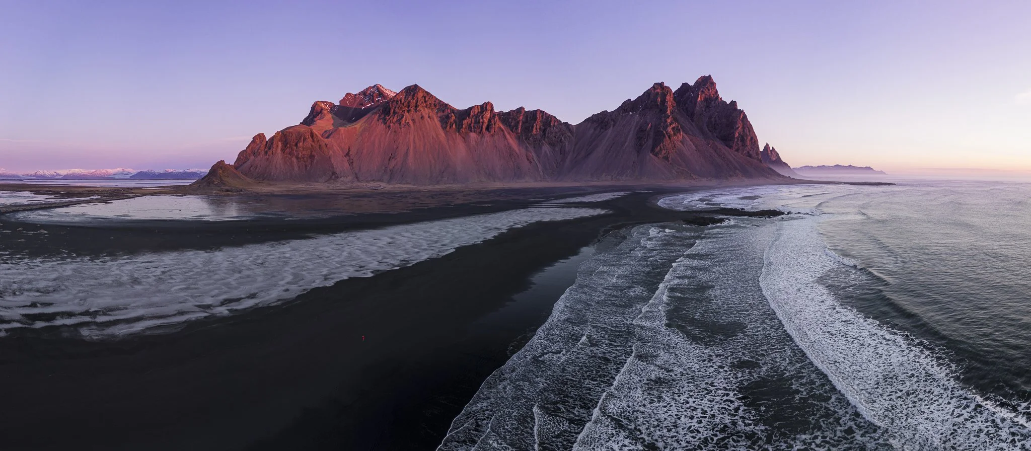 A mountain range with reddish peaks during sunset, next to a black sand beach with ocean waves.