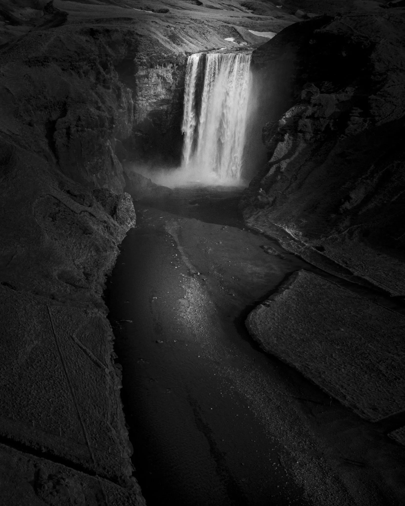 A black and white photograph of a waterfall flowing over rocks into a pool below, surrounded by rugged cliffs.