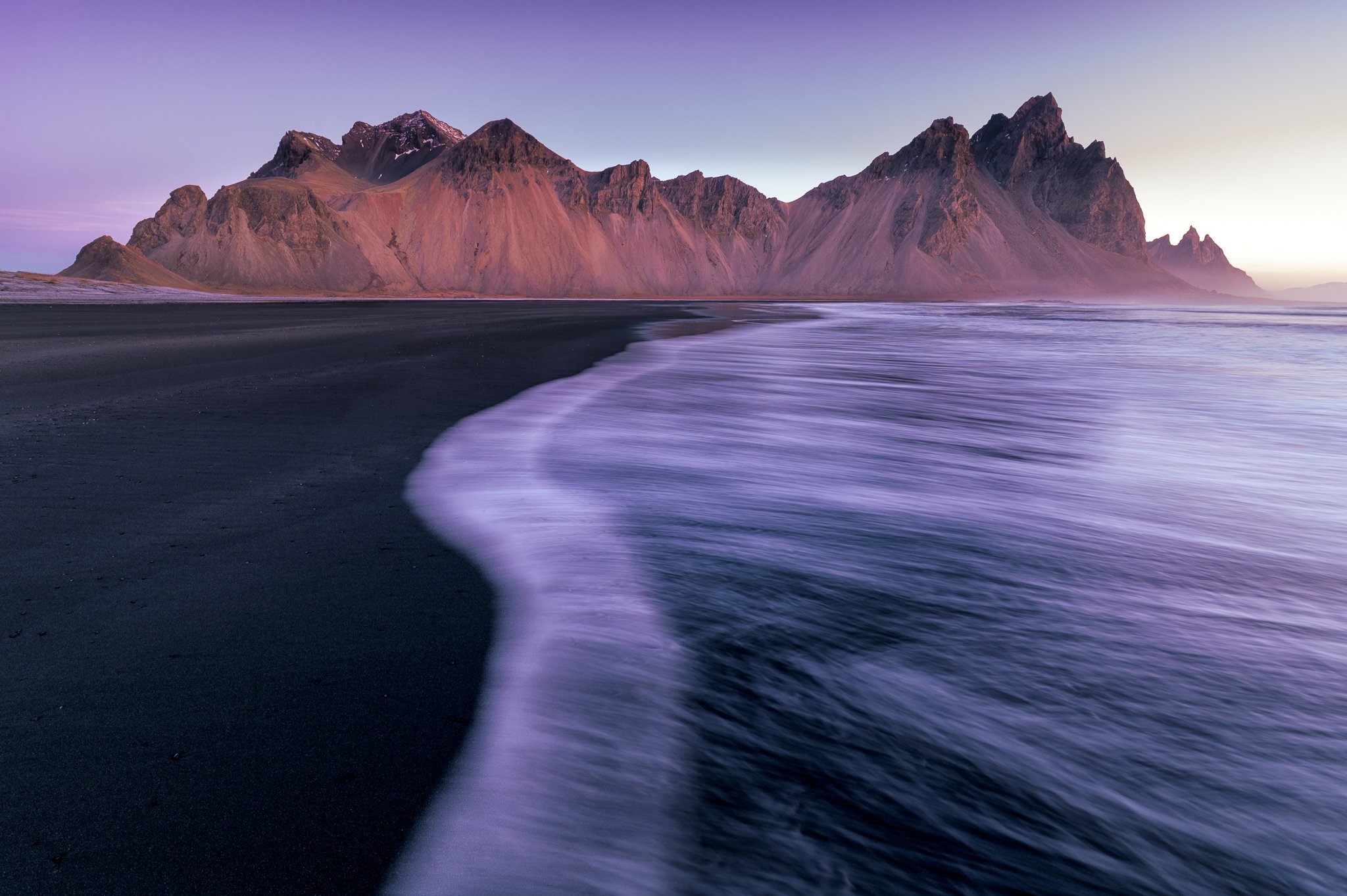A scenic view of black sand beach with mountains in the background and waves washing onto the shore at sunset or sunrise.