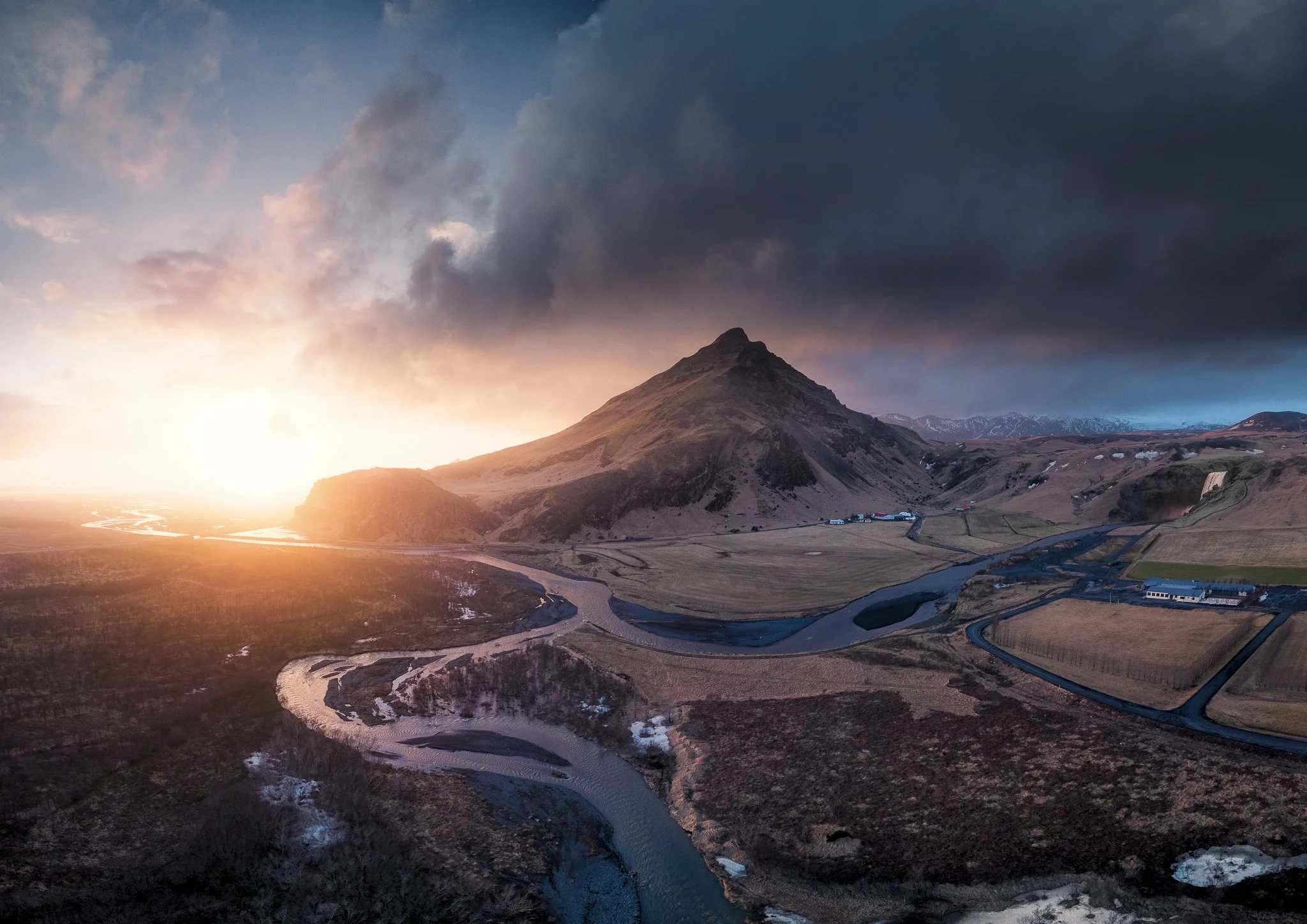 A mountain landscape at sunset with a winding river, small buildings, and dark clouds in the sky.