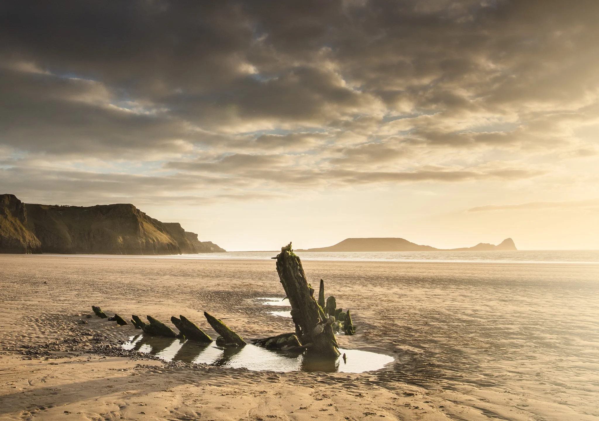 Sunset beach scene with algae-covered wooden posts in the sand, cliffs in the background, and a partly cloudy sky.