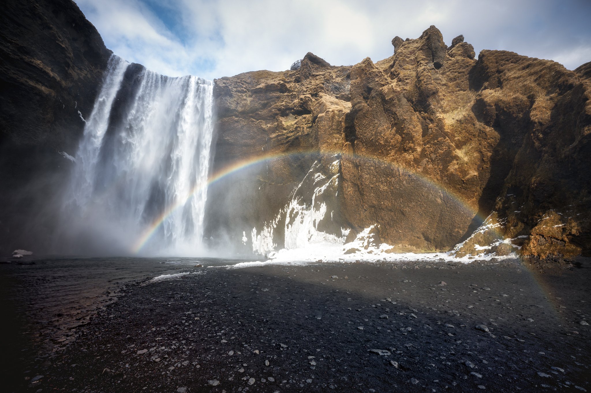 Waterfall surrounded by rocky cliffs with a rainbow arcing across the spray and a cloudy sky above.