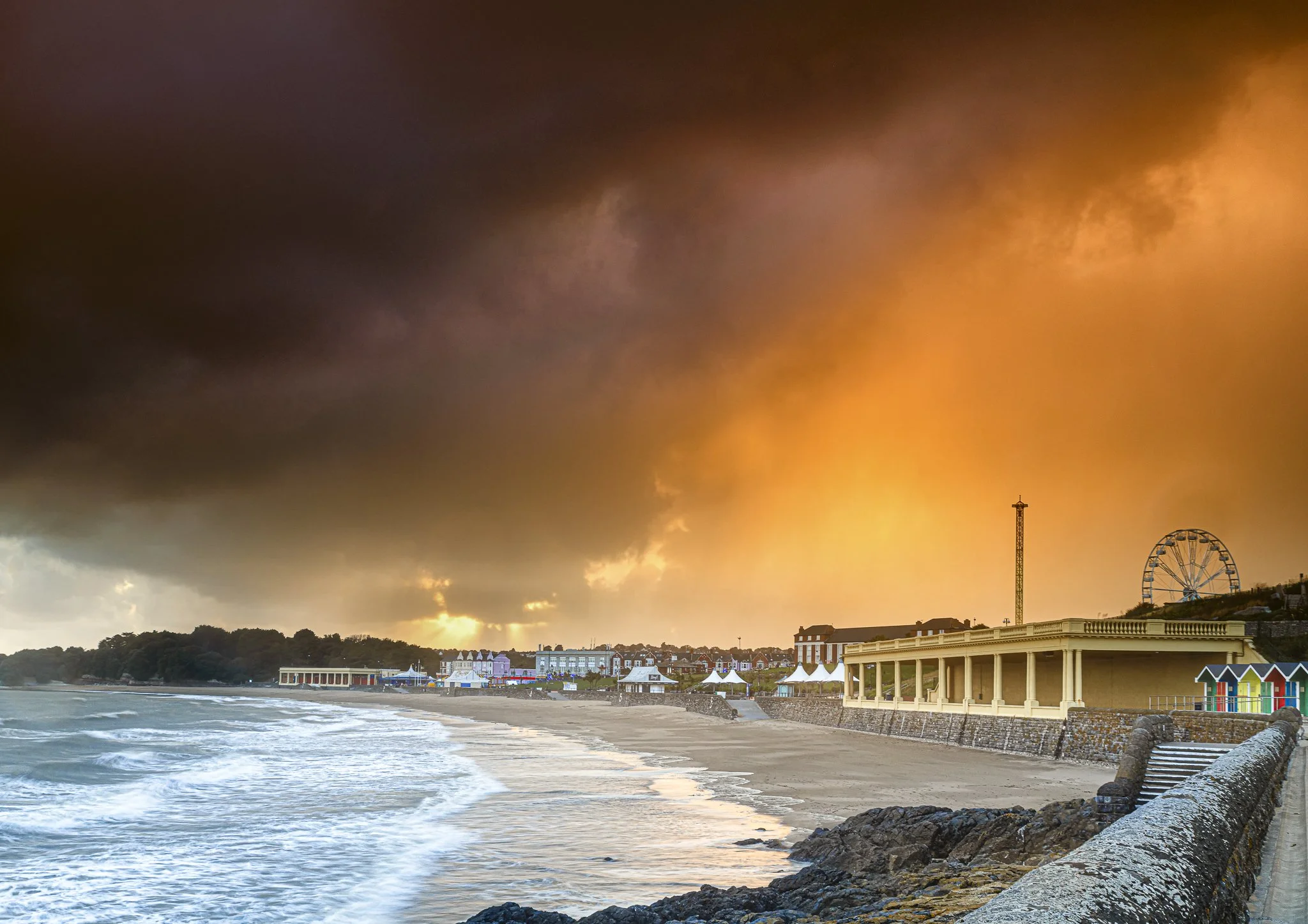 Beach with European-style buildings, a Ferris wheel, and a stormy orange and dark sky.