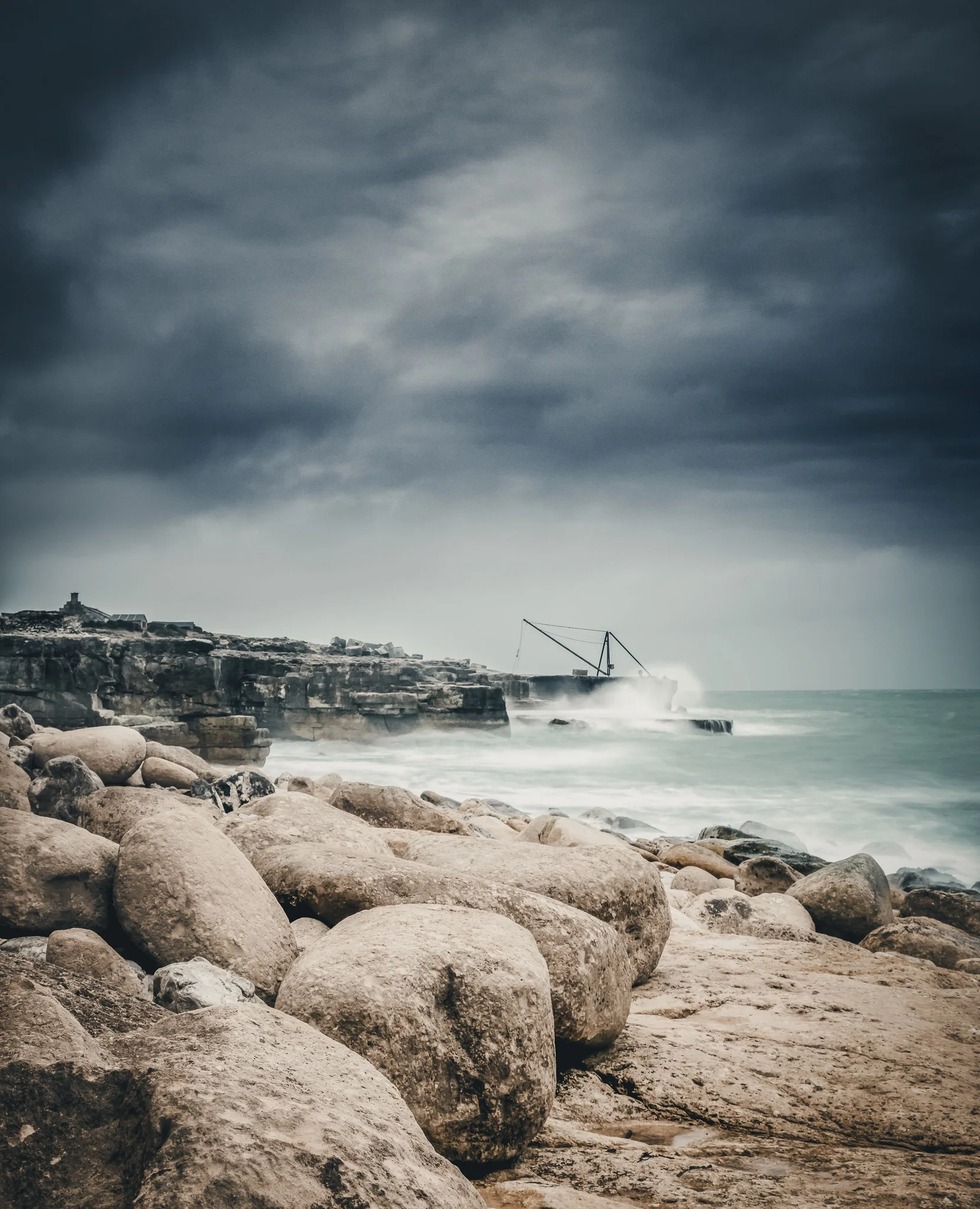 Rocky beach with large stones in the foreground, overcast sky, crashing waves, and a partially broken pier in the distance.