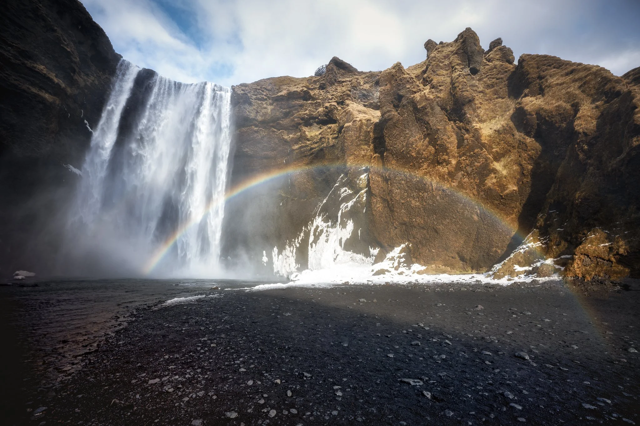 Waterfall inside a canyon with a rainbow arched across the scene, dark rocky ground in the foreground.